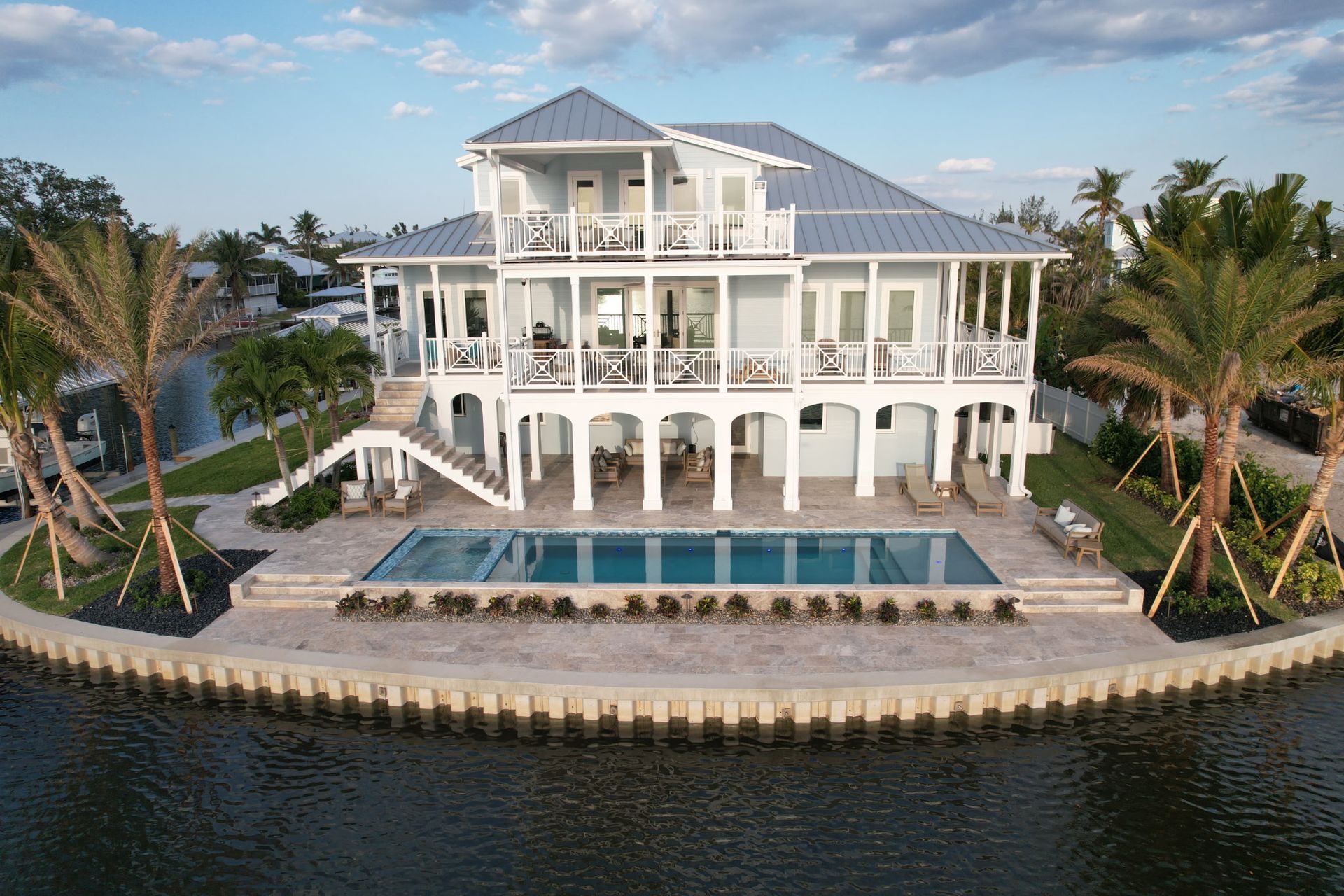 Two-story blue and white house on water, with a pool, balconies, and palm trees.