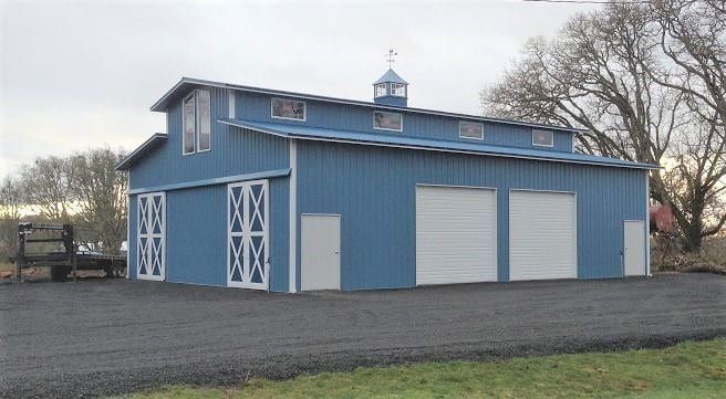 Blue barn with white doors and garage doors, on a gravel driveway. Overcast sky.