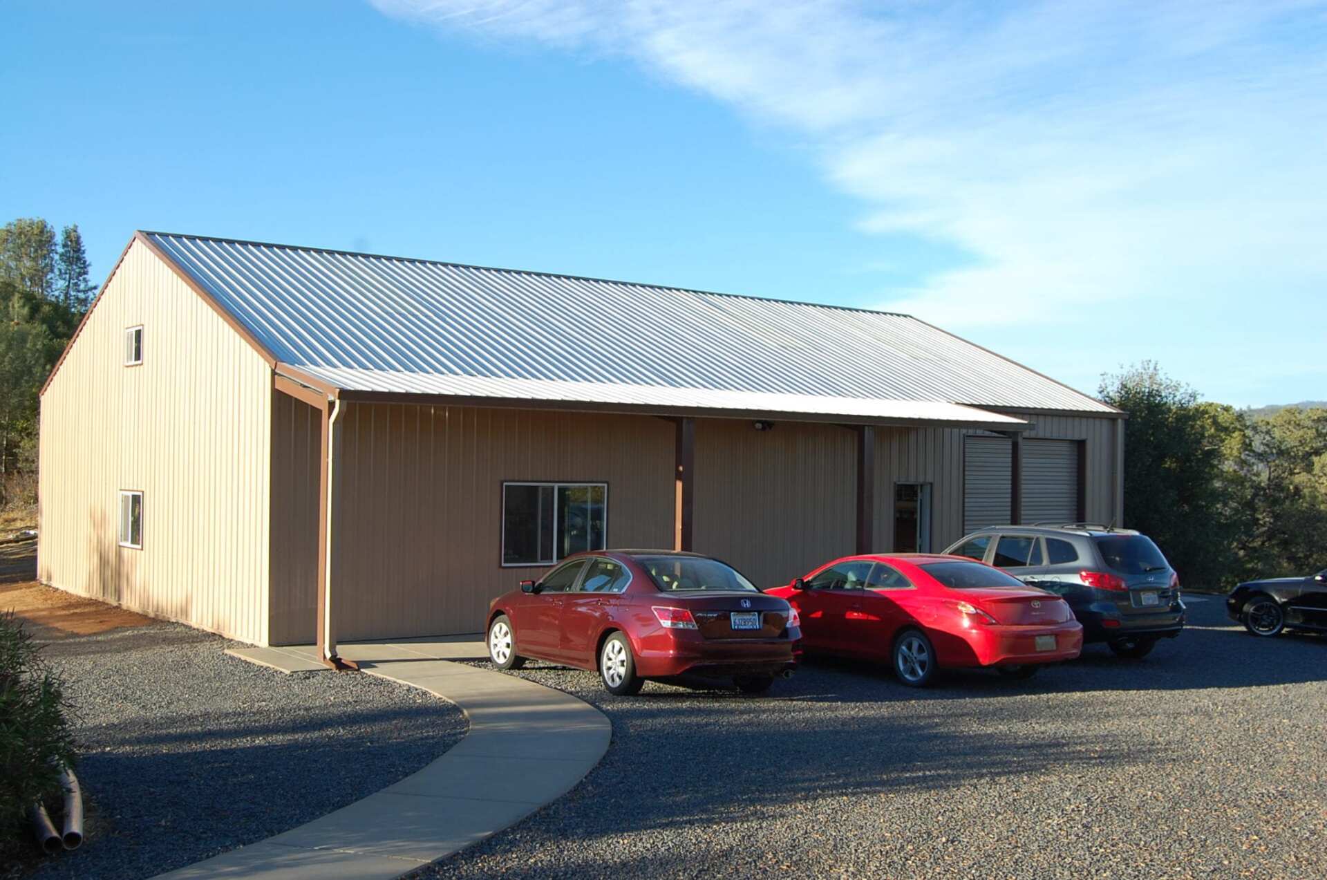 Tan metal building with corrugated silver roof; cars parked in front on gravel.