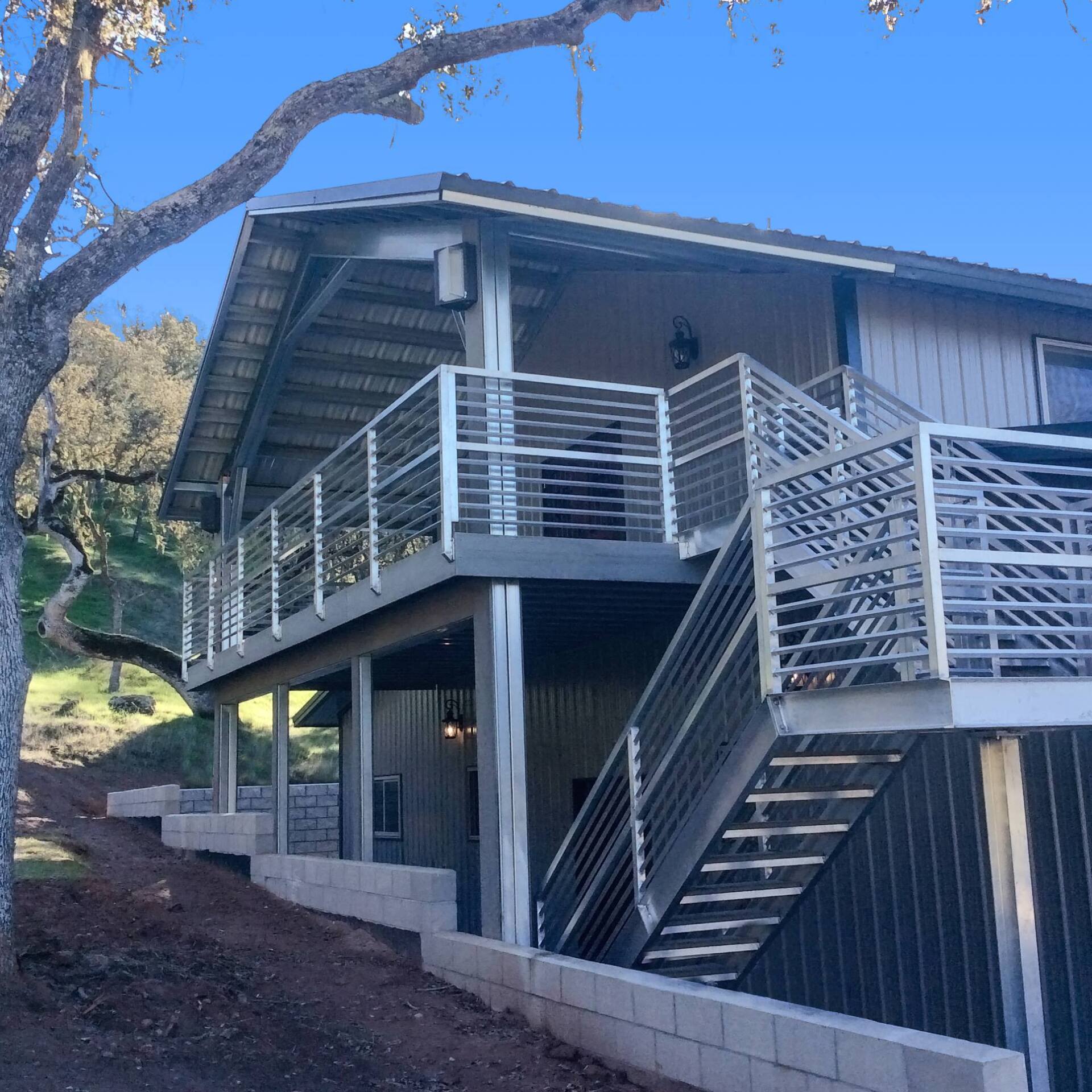 Two-story modern home with steel railings, stairs, and a metal roof. Gray siding against a hillside.
