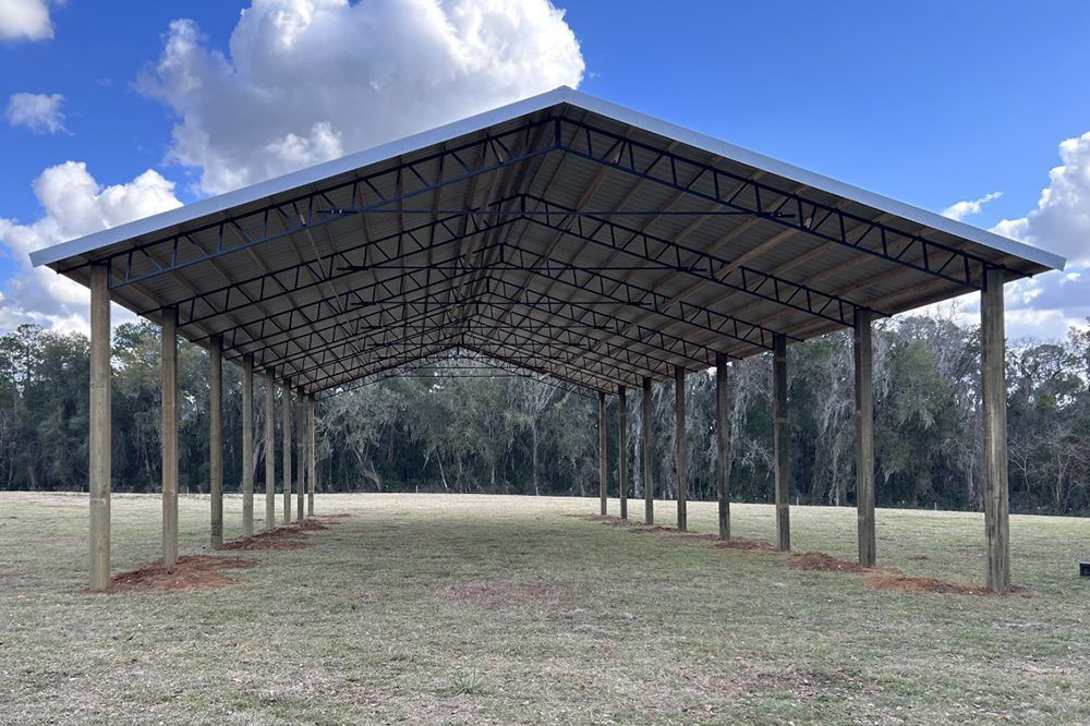 Open-sided metal and wood barn-style structure with a sloping roof, set in a grassy field under a blue sky.