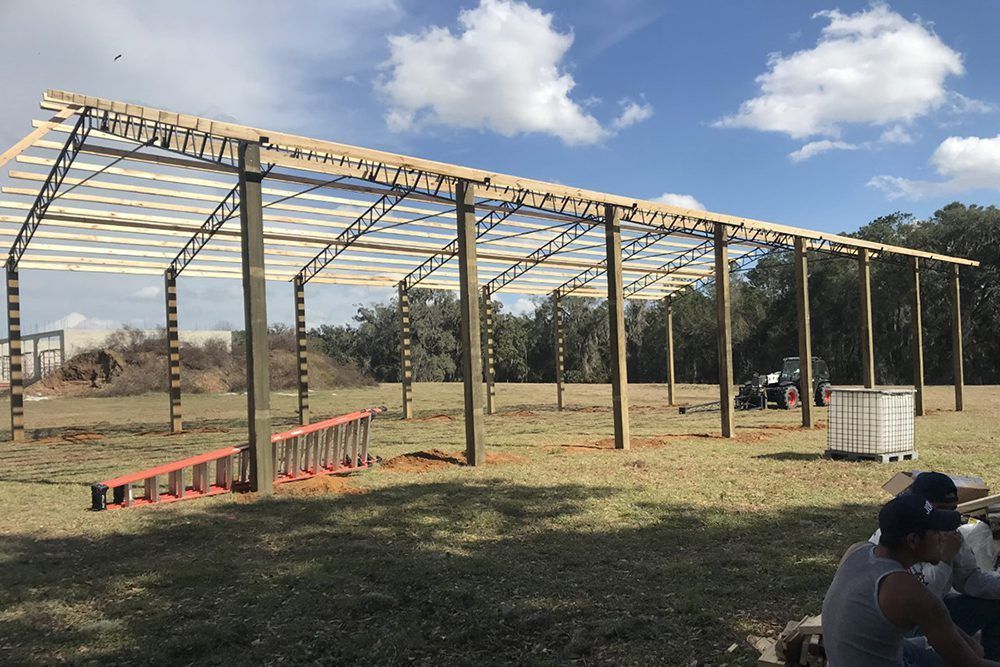 Open-air structure under construction; wooden beams and clear roofing; rural setting with workers and equipment.