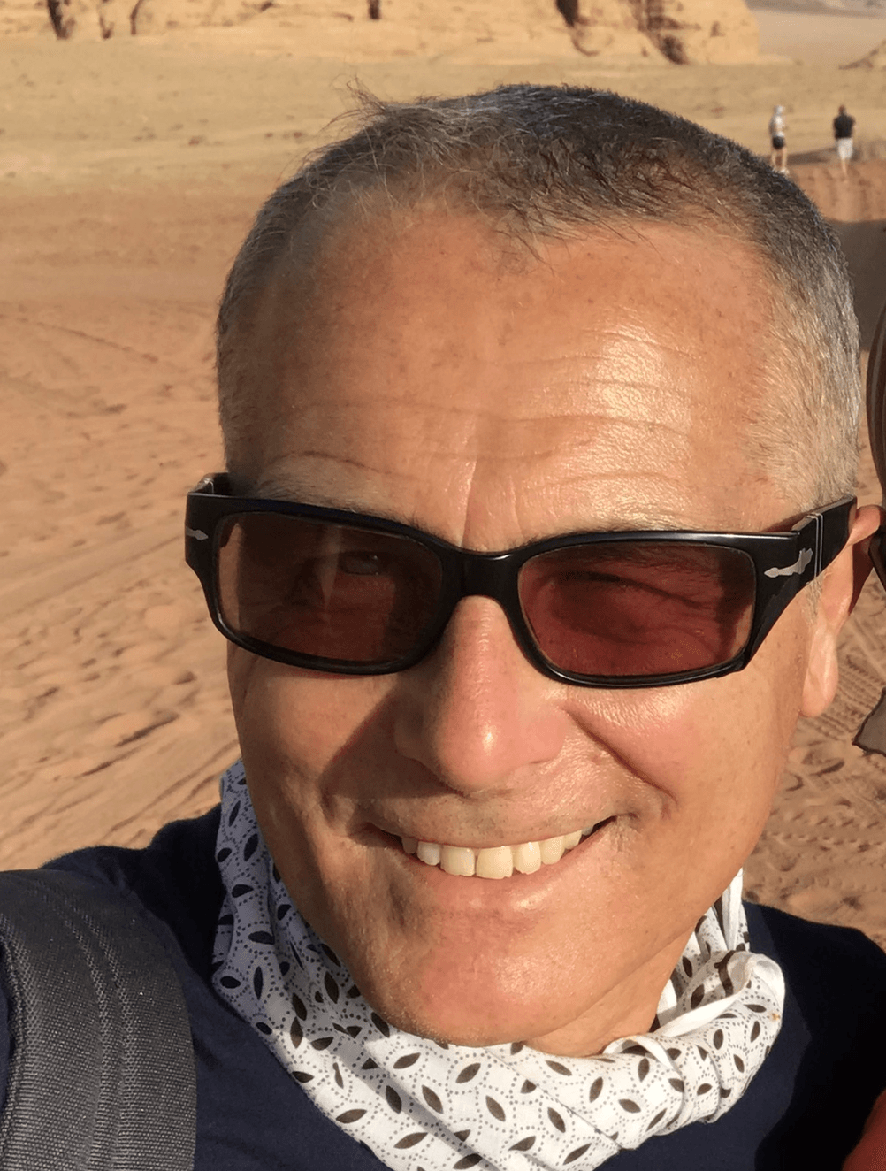 Una persona sorridente con occhiali da sole e una sciarpa a fantasia al collo, in un ambiente desertico sabbioso.