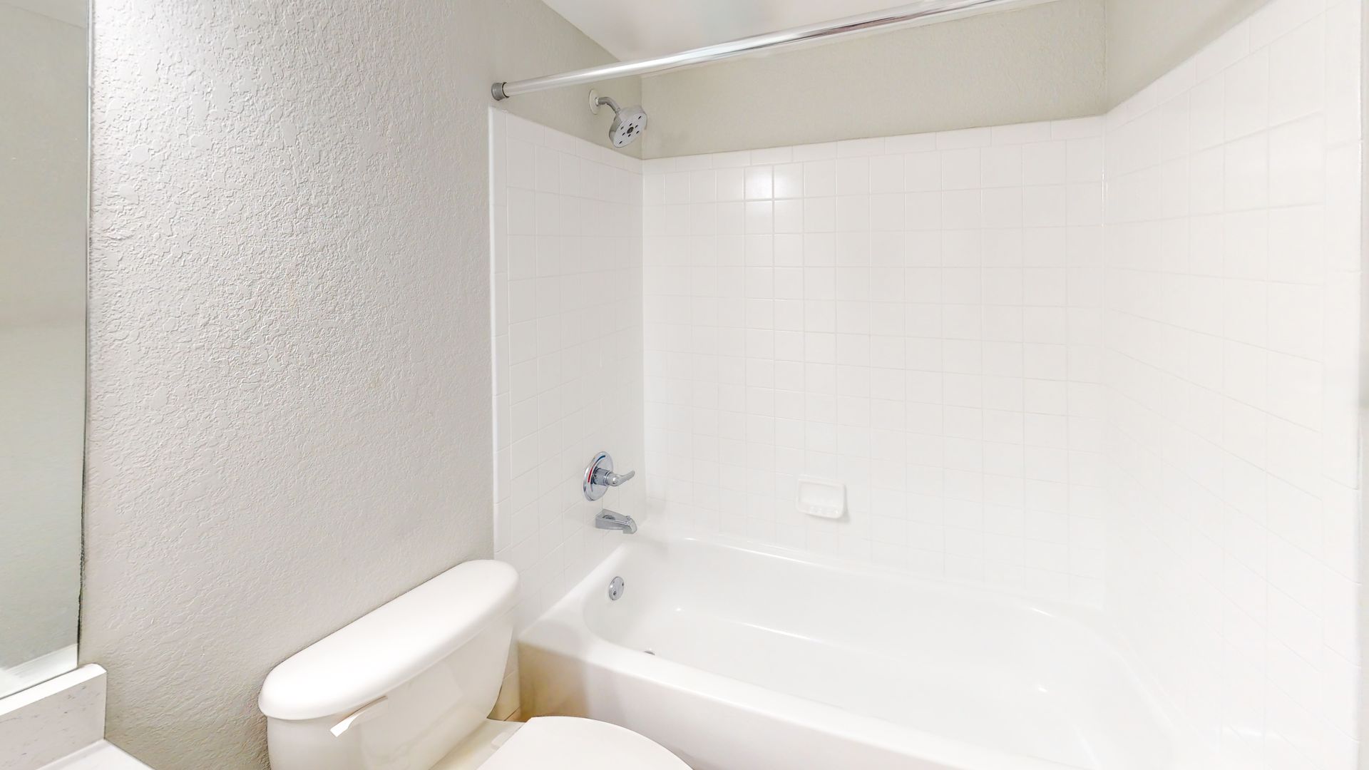 Small bathroom with white tiled tub and shower surround, toilet, and beige textured wall.