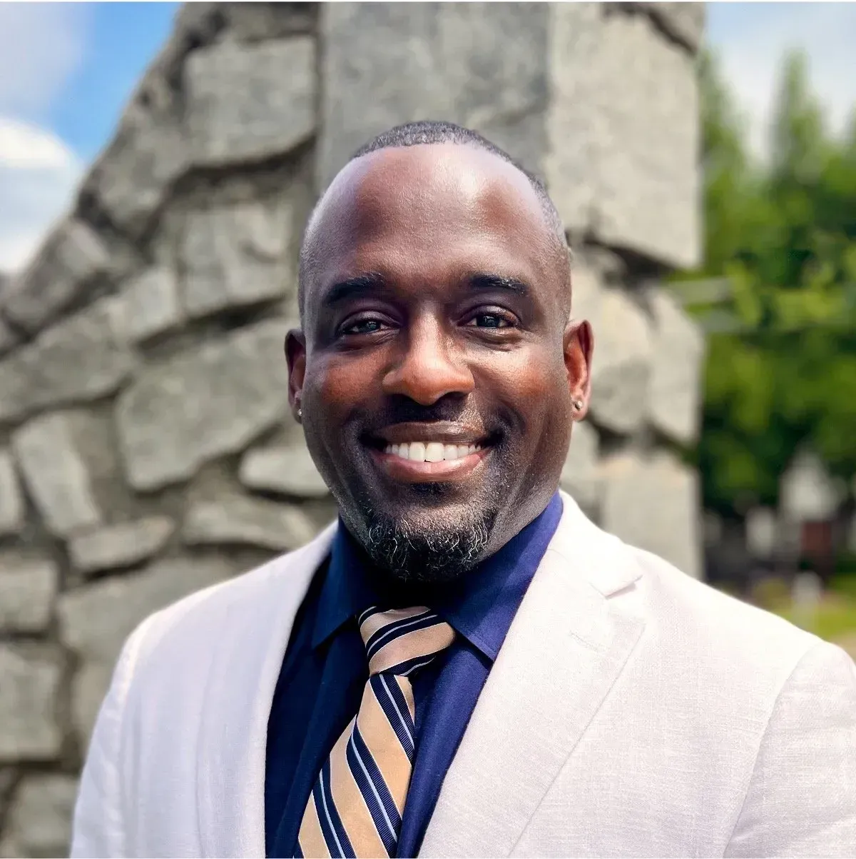Smiling man in a light suit and striped tie standing outdoors before a stone wall