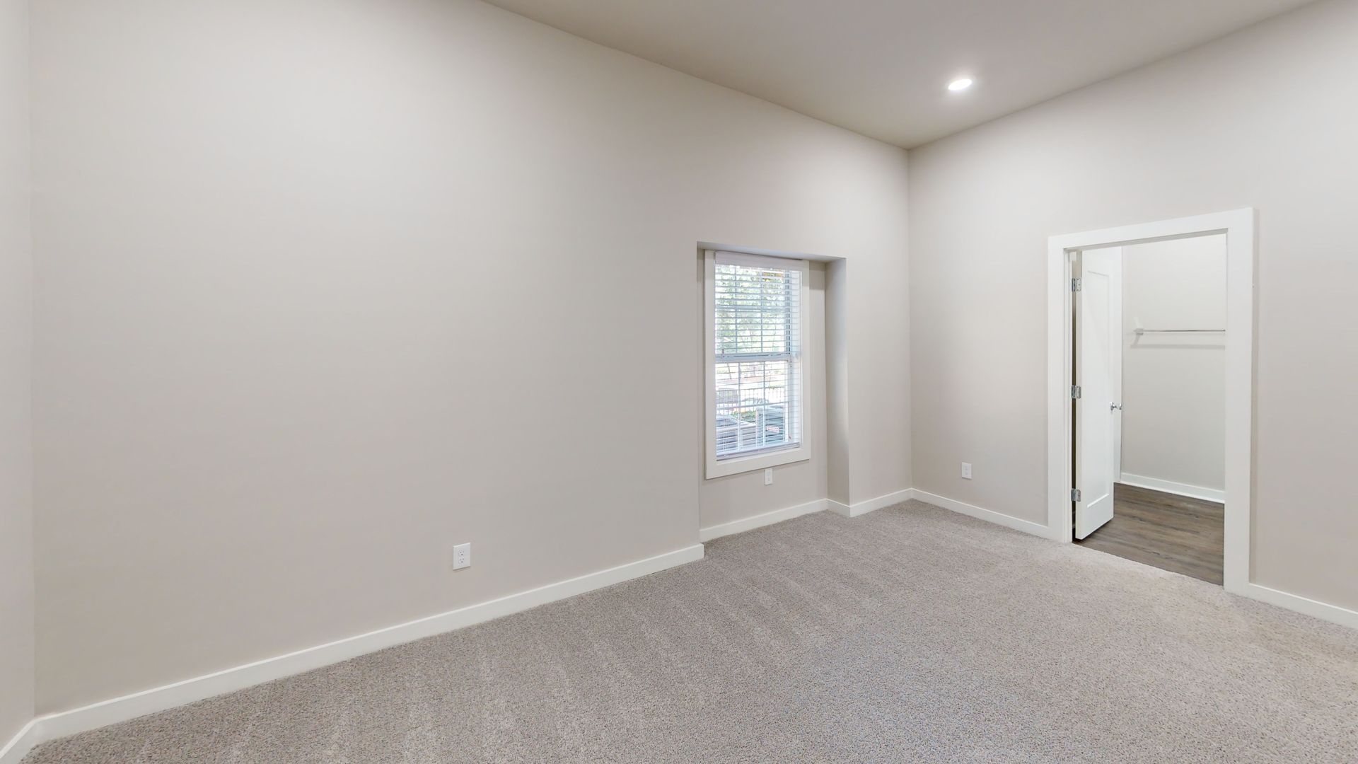 Empty carpeted bedroom with light gray walls, a window, recessed lighting, and an open closet door