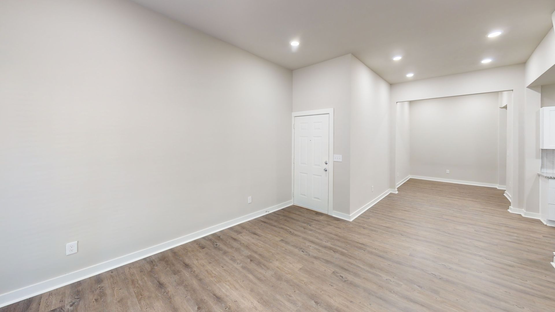 Empty modern room with light gray walls, wood flooring, recessed lights, and a white door