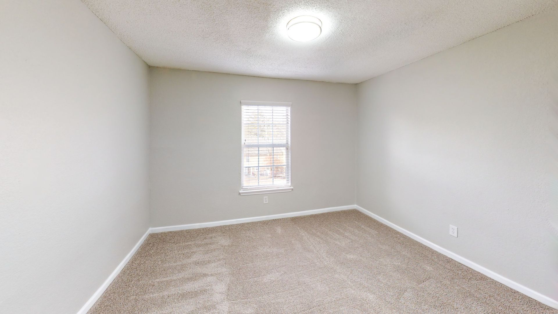 Empty beige bedroom with white walls, carpet, a small window, and a ceiling light.