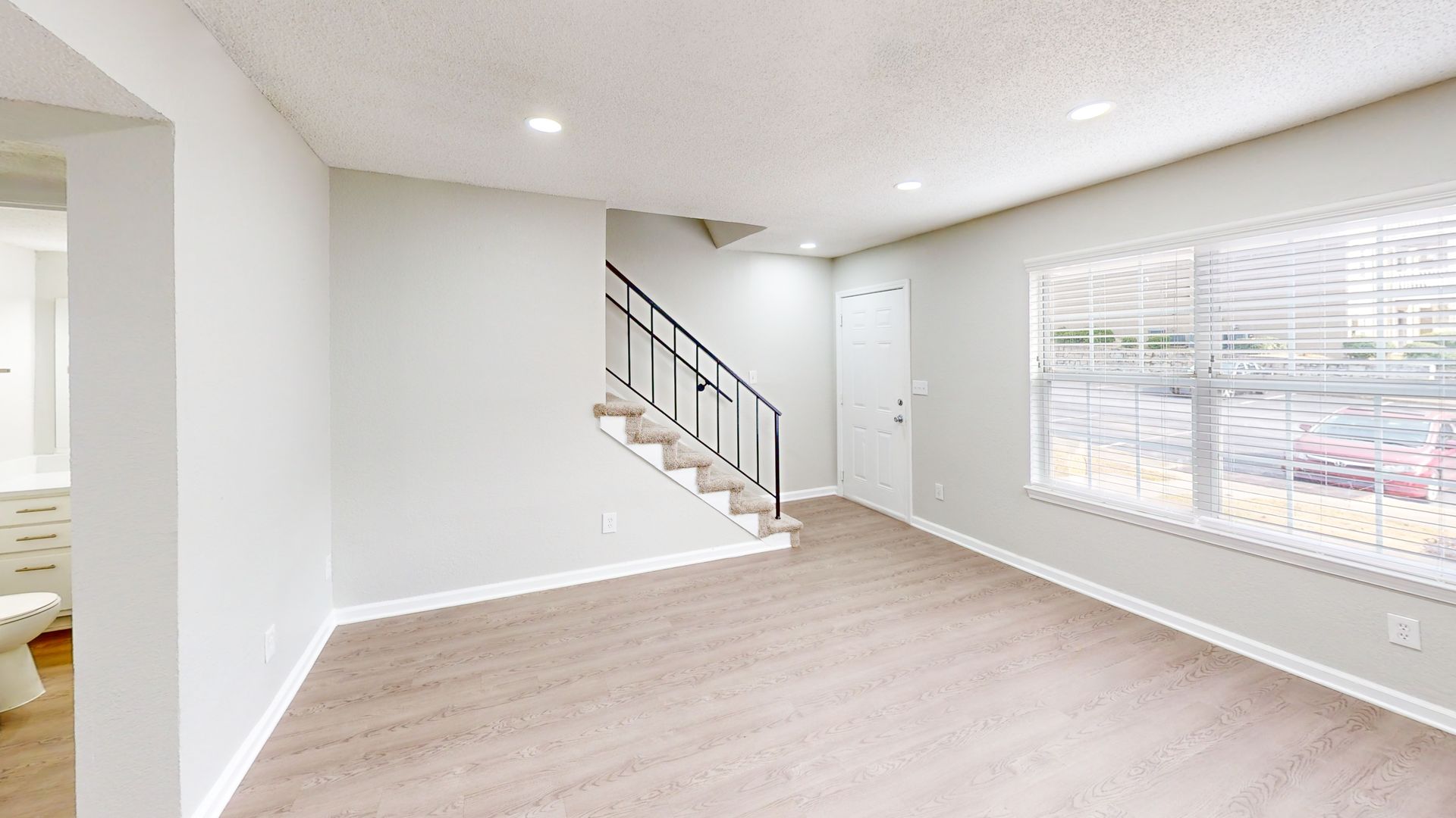 Empty bright living room with light gray walls, wood floors, stairs, and large windows.