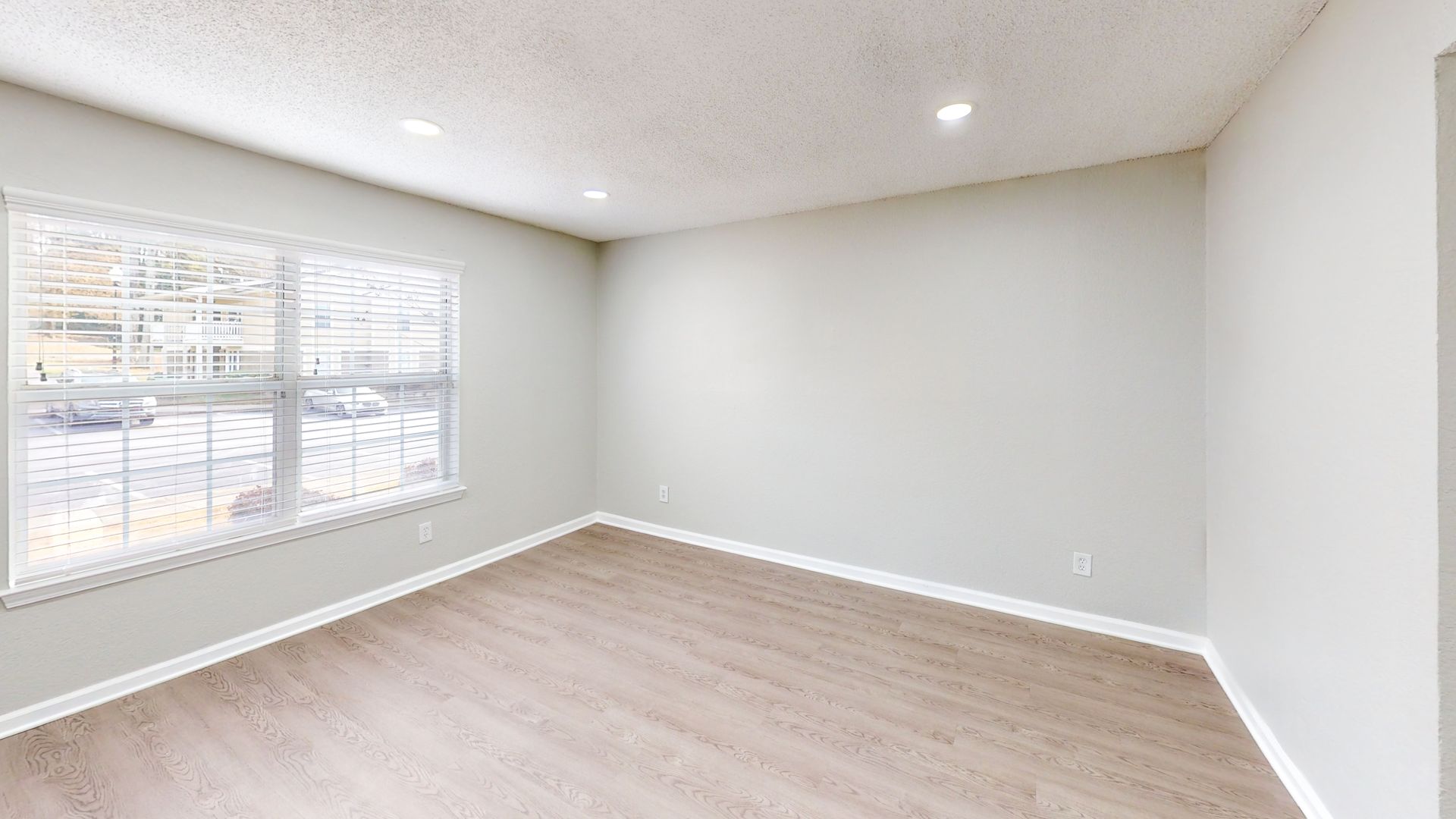 Empty bright room with white walls, light wood floor, recessed lighting, and a large window with blinds.