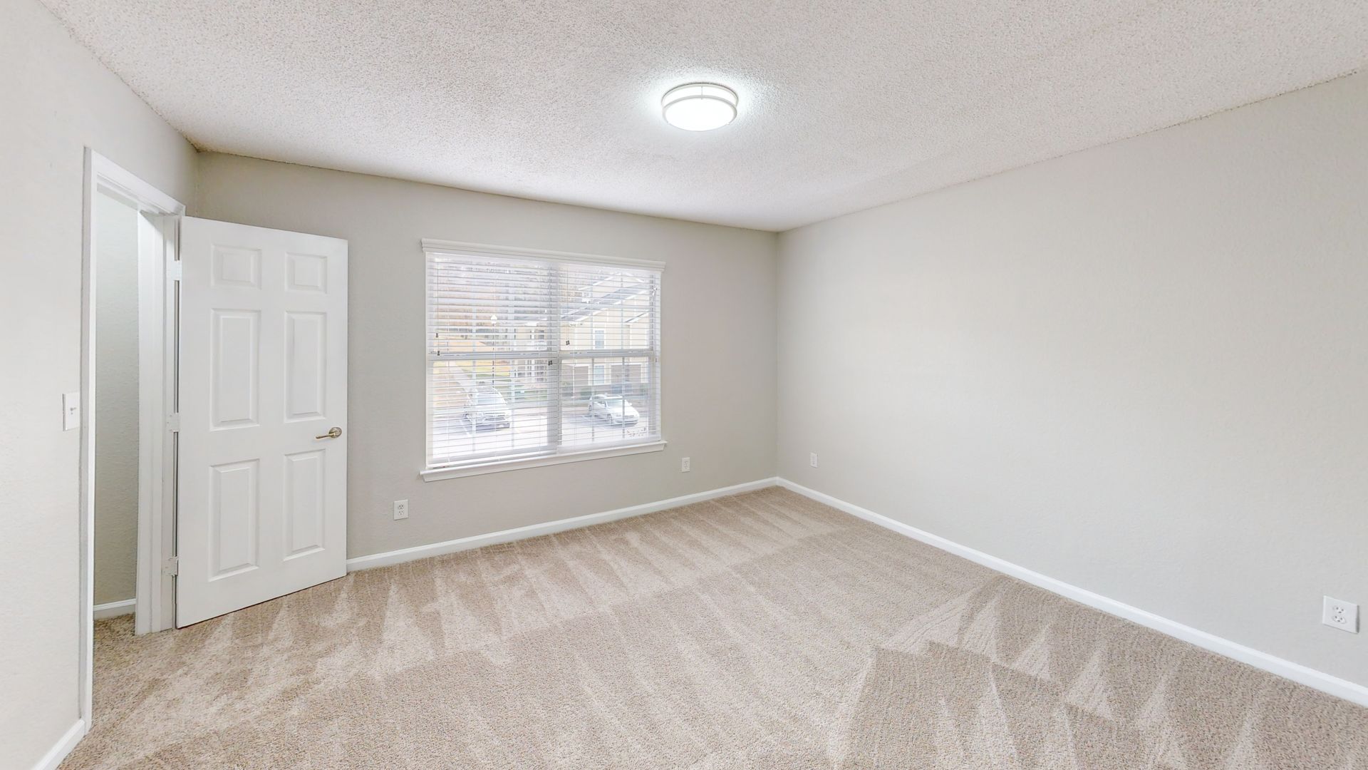 Empty beige bedroom with carpet, white walls, closet doors, and a window.