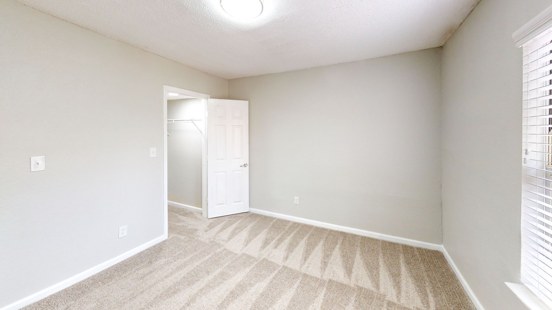 Empty beige bedroom with carpet, open closet, and a window with blinds.