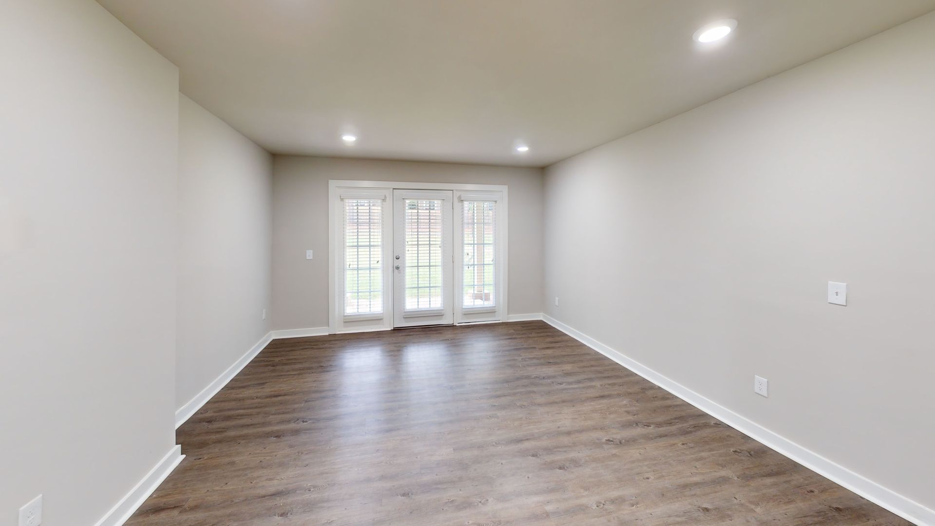 Empty modern living room with gray walls, wood floors, recessed lights, and French doors at the far end