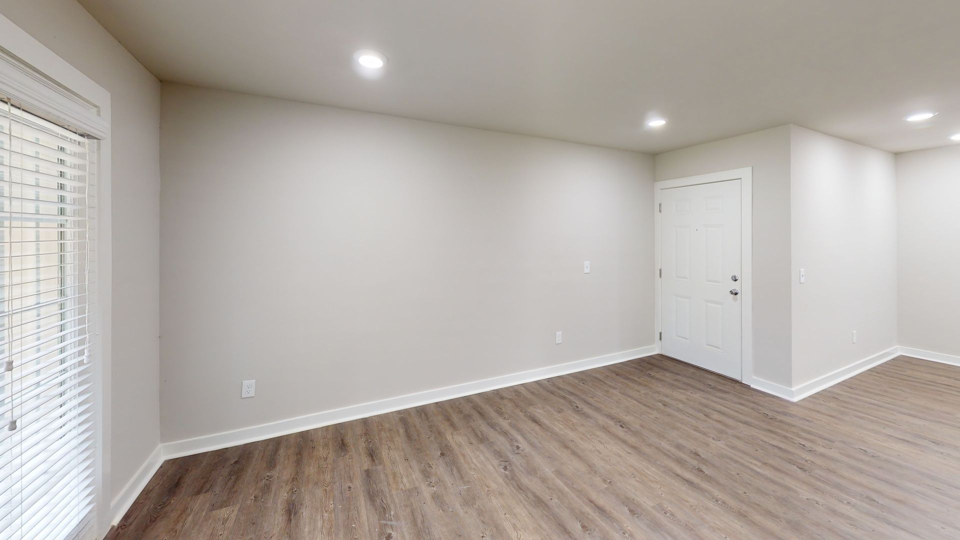 Empty modern living room with light gray walls, wood flooring, and recessed ceiling lights.