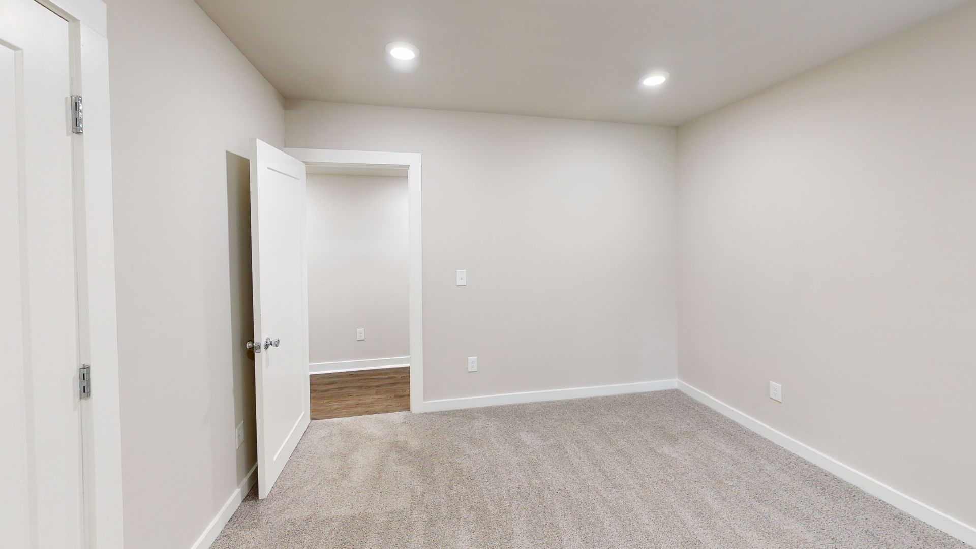 Empty carpeted bedroom with white walls, recessed ceiling lights, and an open door to a hallway