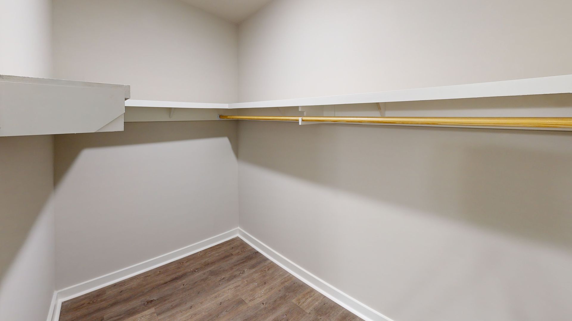 Empty corner closet with white walls, wood floor, and a single shelf along the back and side walls