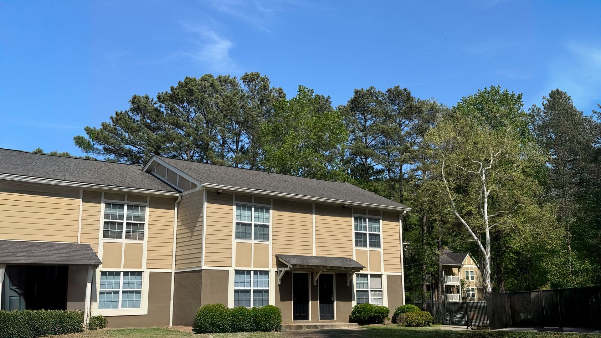 Two-story beige apartment building with dark roof and tall trees under a clear blue sky