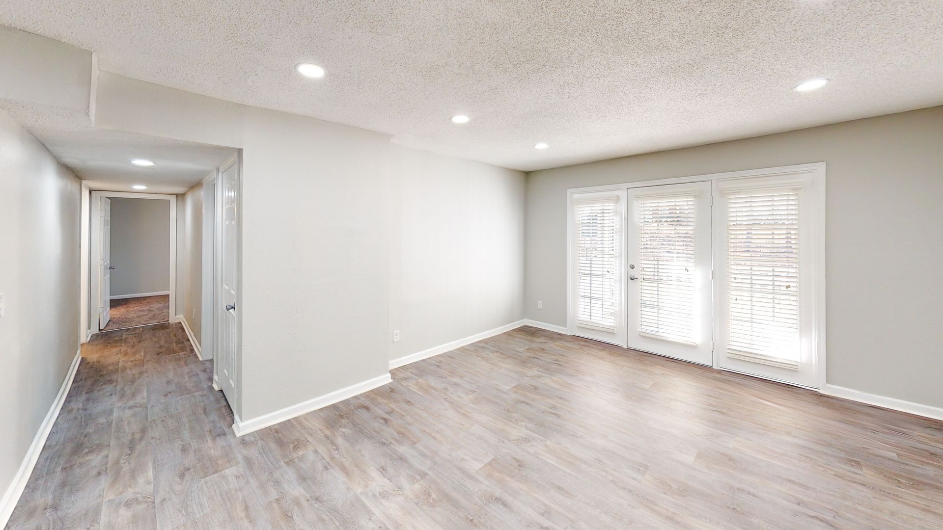 Empty modern room with light wood floors, white walls, recessed lighting, and balcony doors.