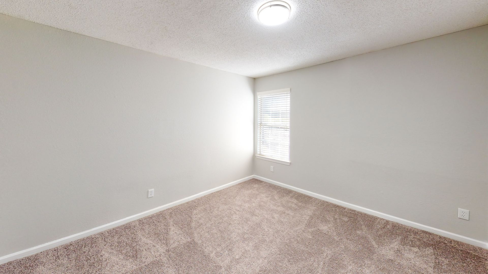 Empty beige carpeted room with white walls, a small window, and ceiling light.