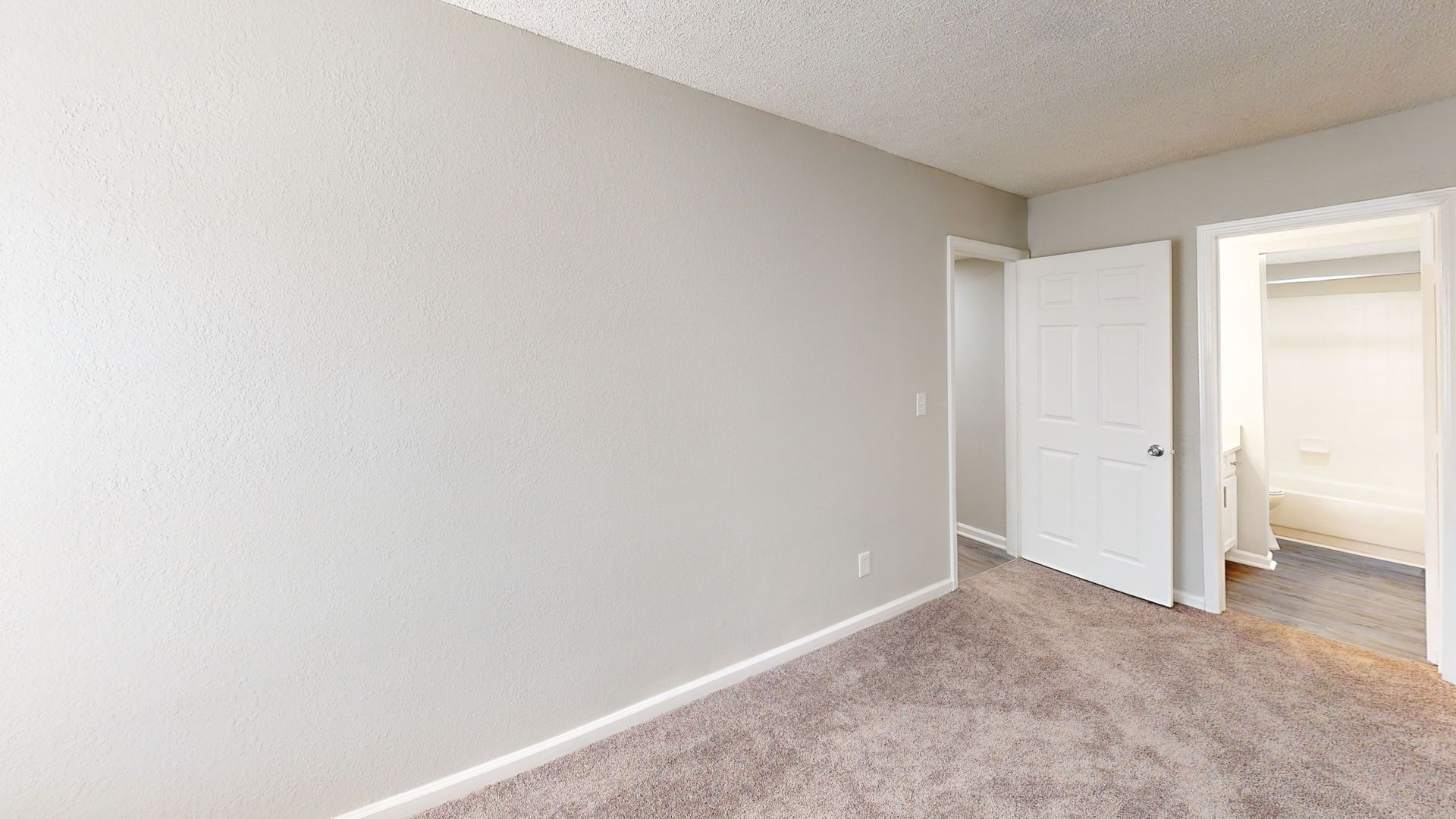 Empty beige bedroom with carpeted floor and open door to a bright hallway