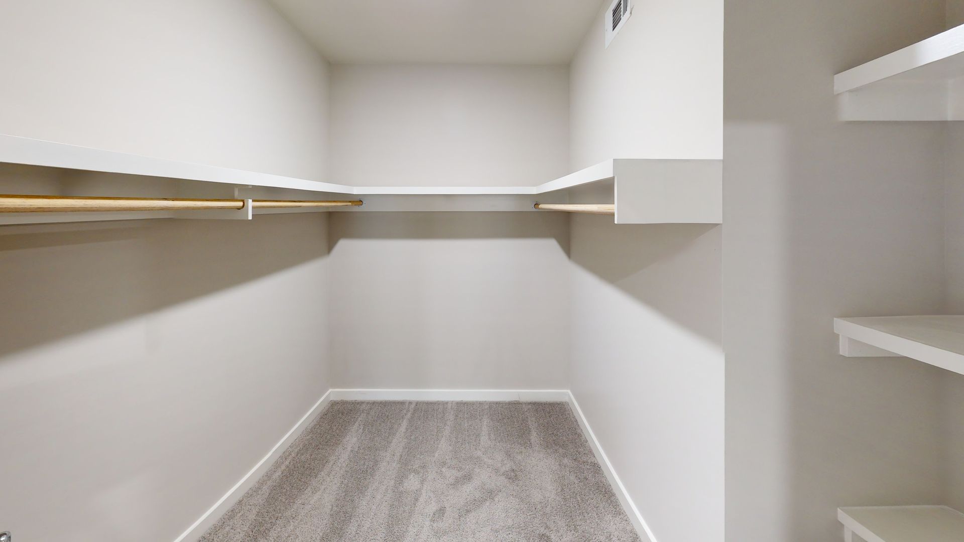 Empty walk-in closet with white walls, wood shelves, and gray carpet flooring.