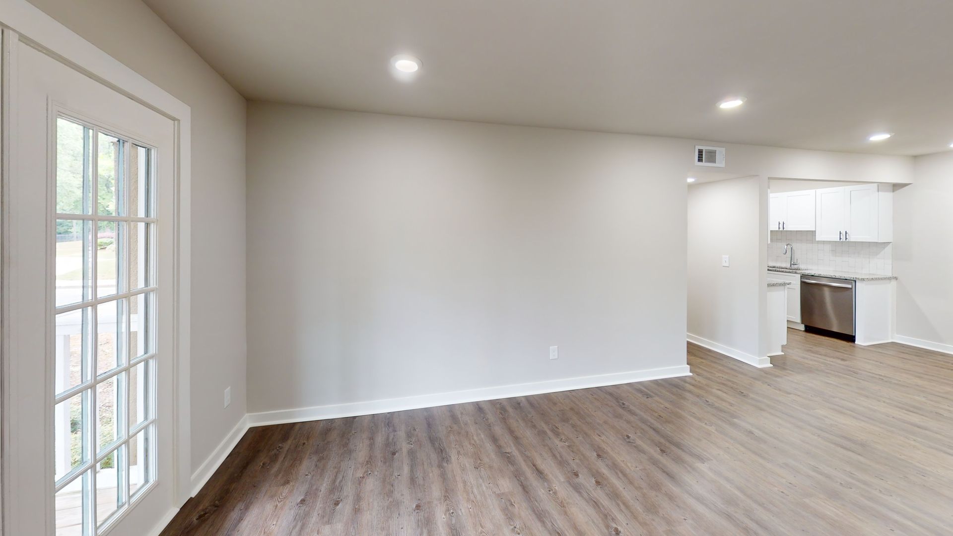 Empty living room with wood floors, gray walls, and an open view into a white kitchen.