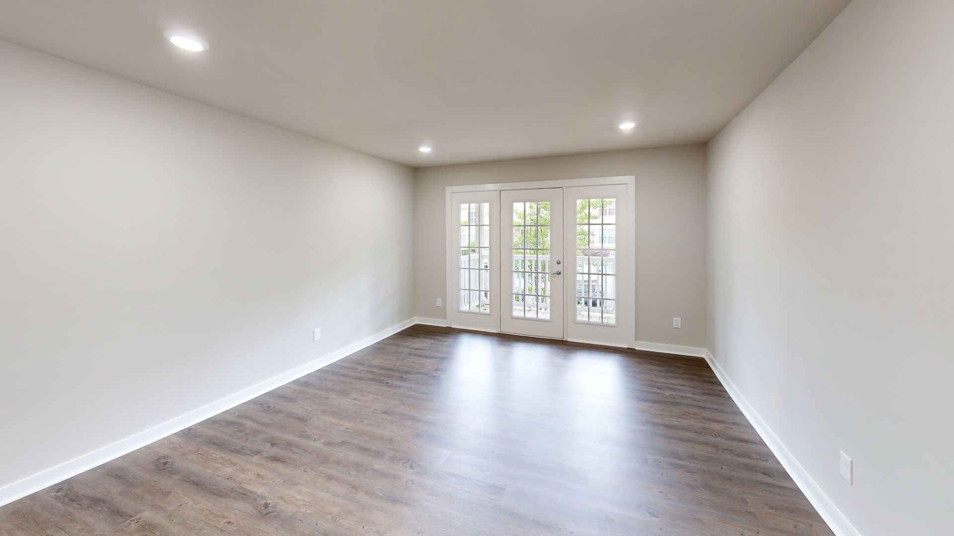 Empty living room with white walls, wood floors, recessed lights, and glass double doors to a patio.