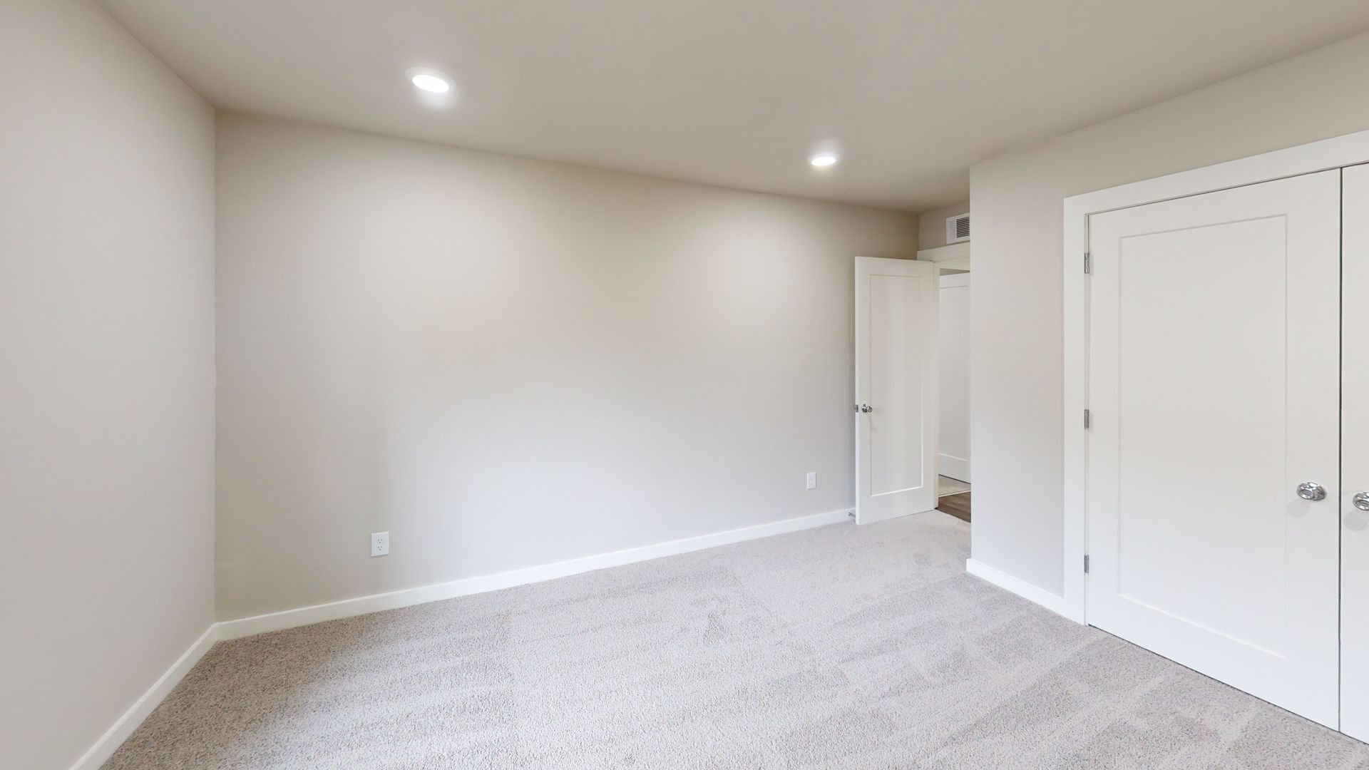 Empty beige bedroom with carpet, white doors, and a hallway visible in the back