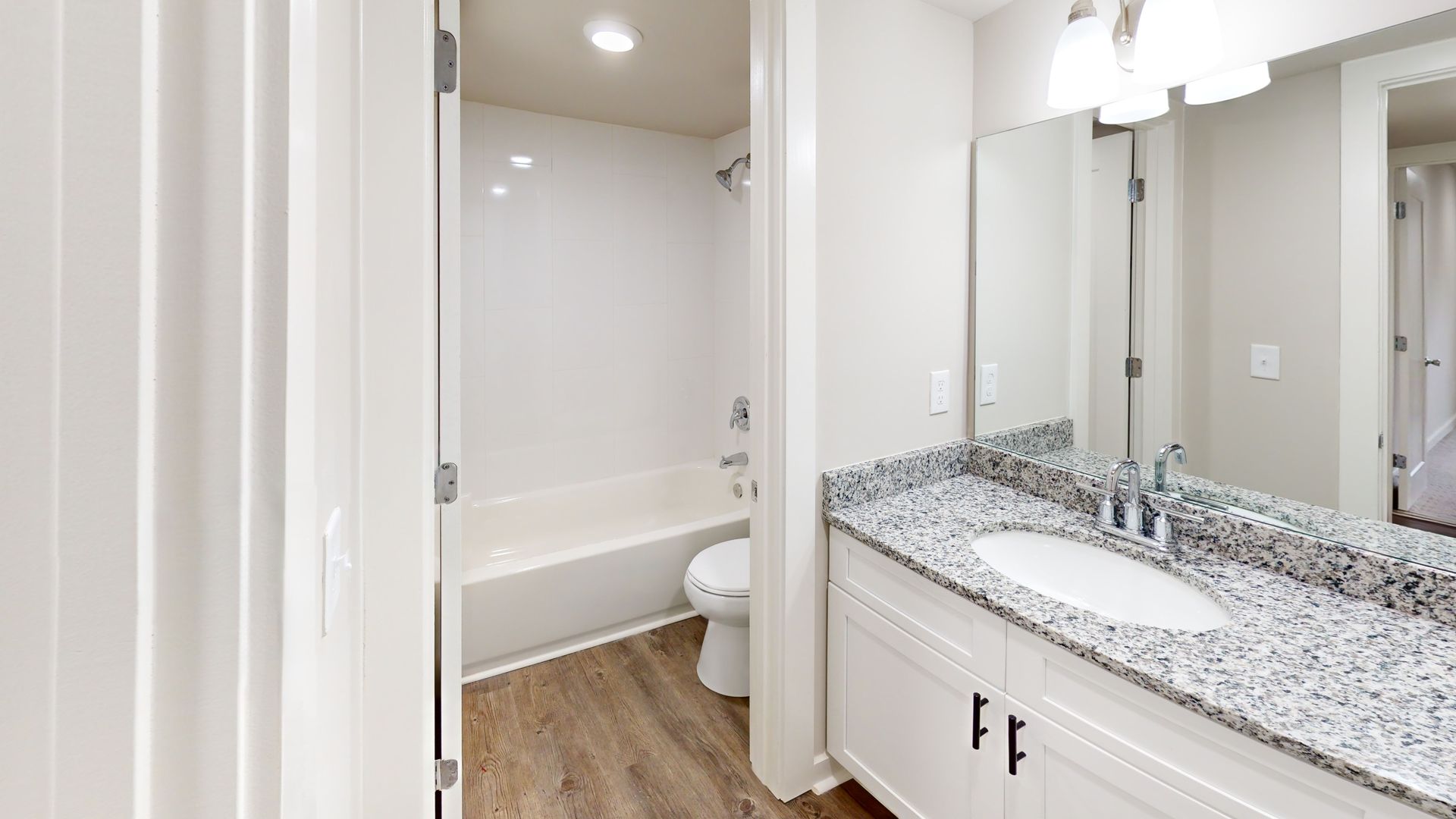 Bright bathroom with double vanity, large mirror, and doorway to a tub and toilet area with wood-look floor.