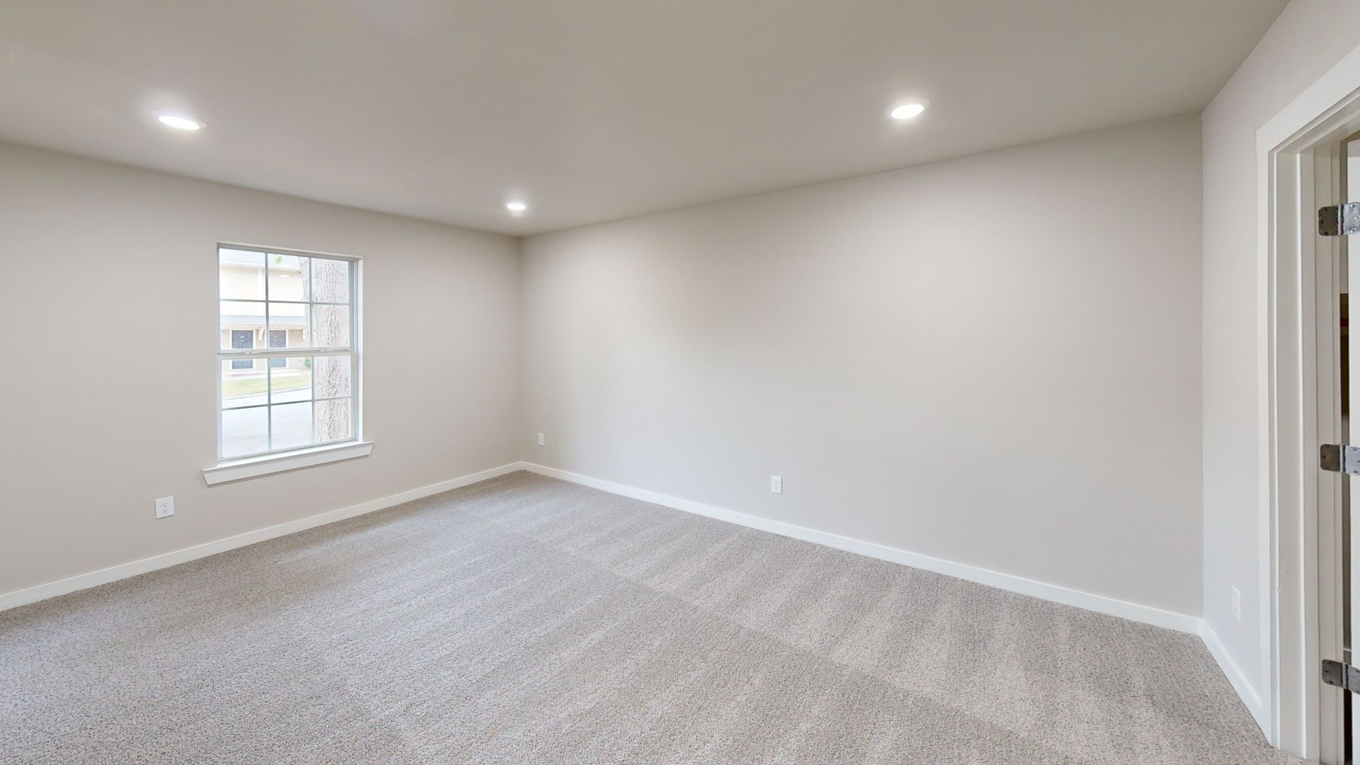 Empty carpeted bedroom with white walls, a window, recessed lighting, and a doorway on the right