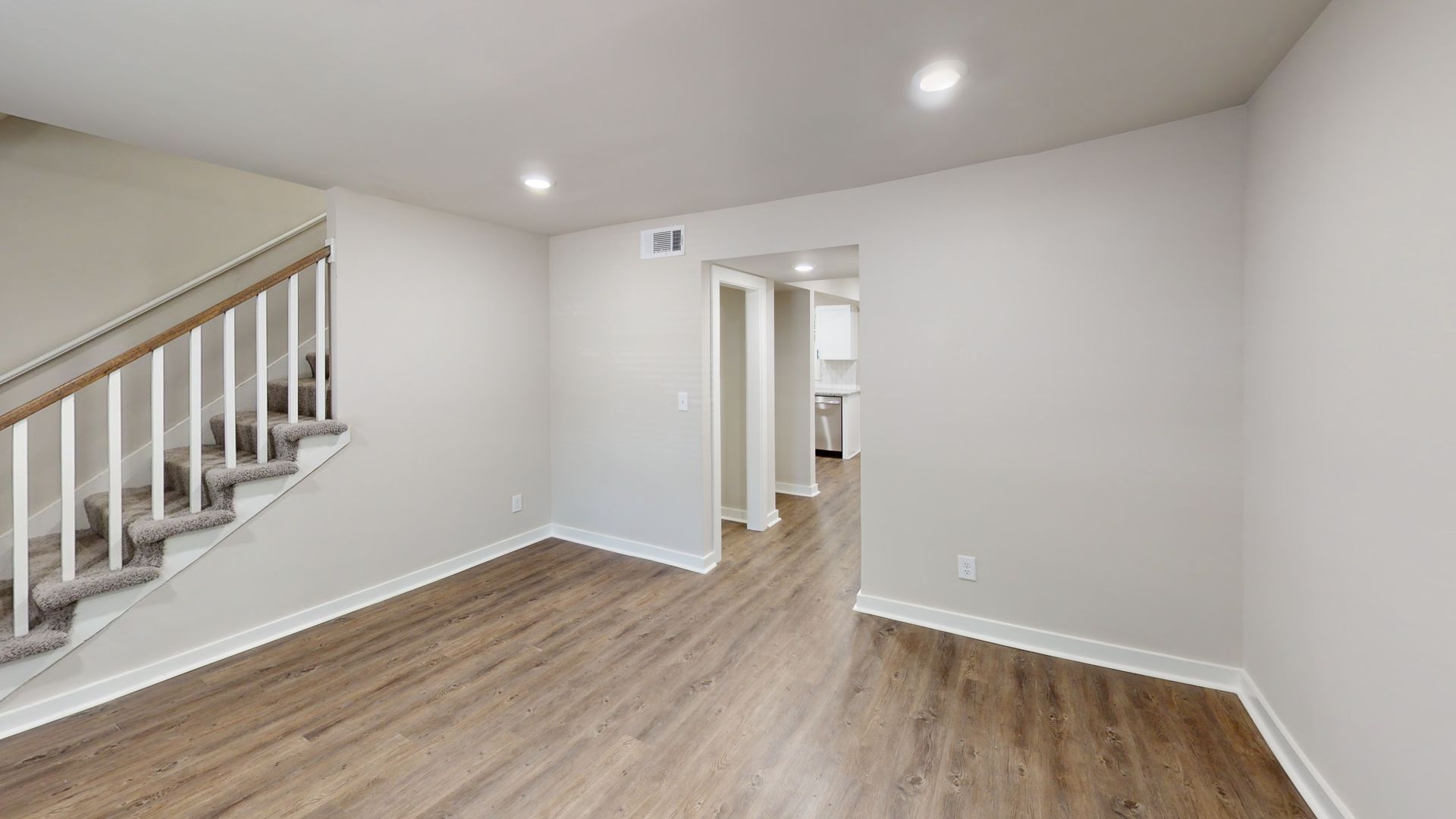 Empty living room with light gray walls, wood floors, and a staircase on the left.
