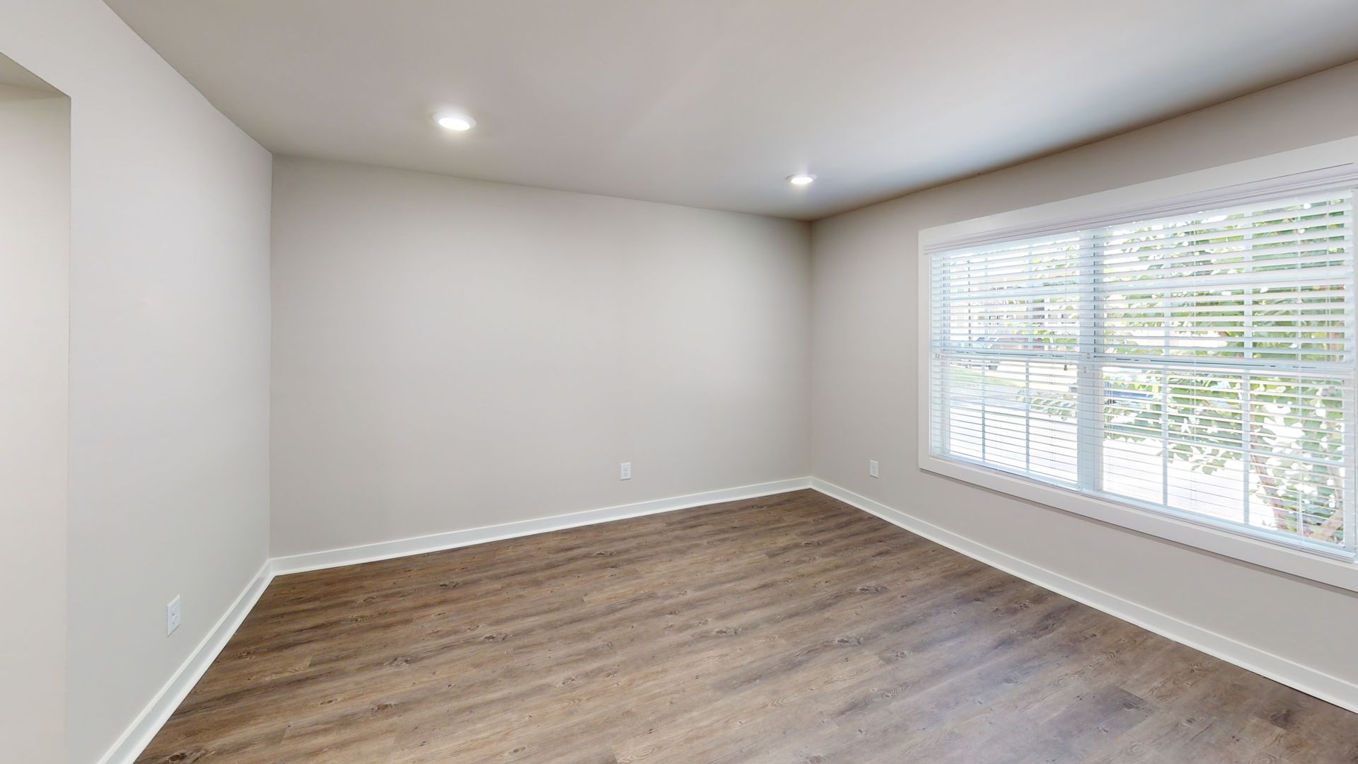 Empty modern bedroom with gray walls, wood flooring, and a large window with blinds.