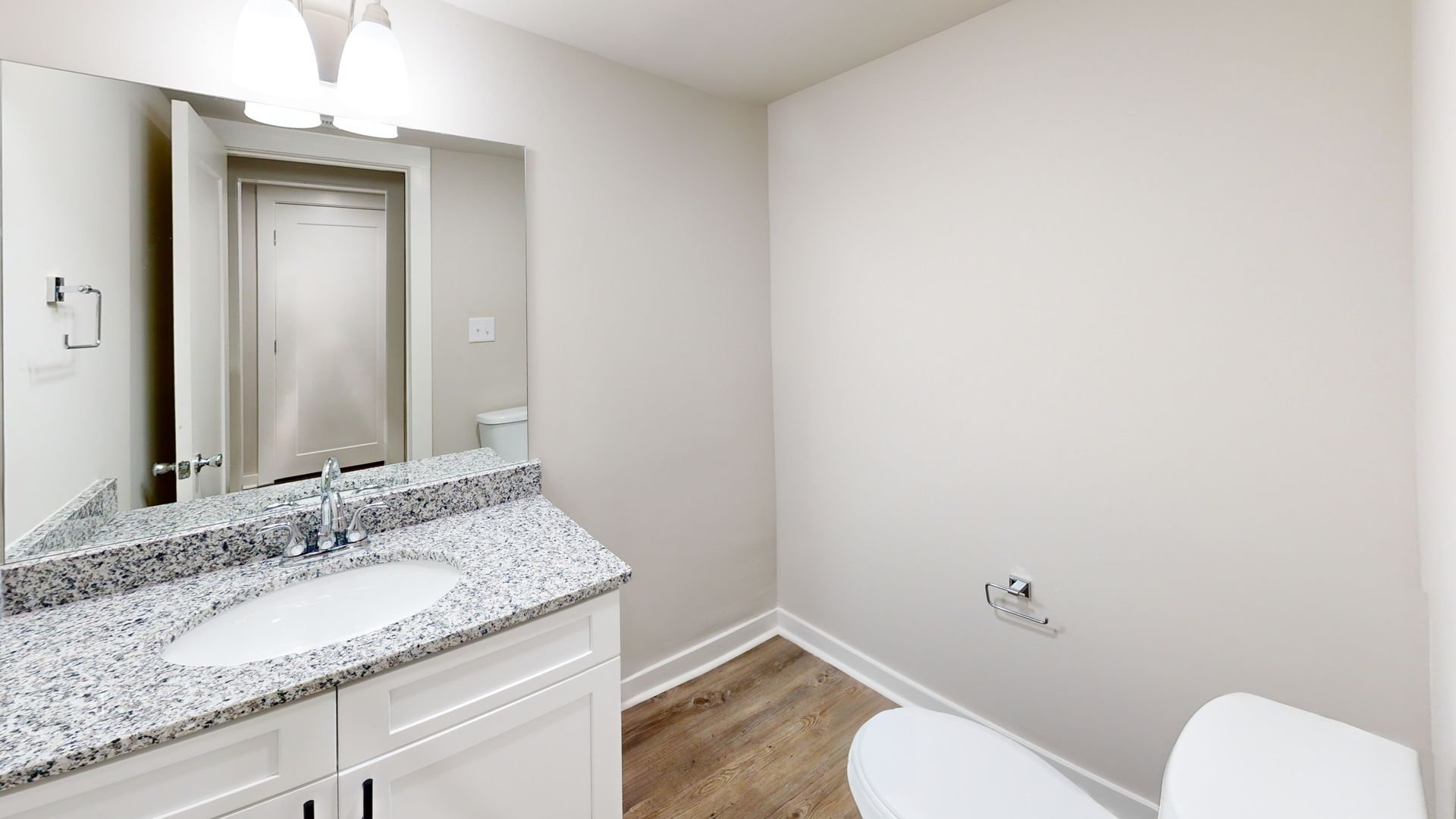 Bathroom vanity with granite countertop, mirror, and toilet in a small white room.