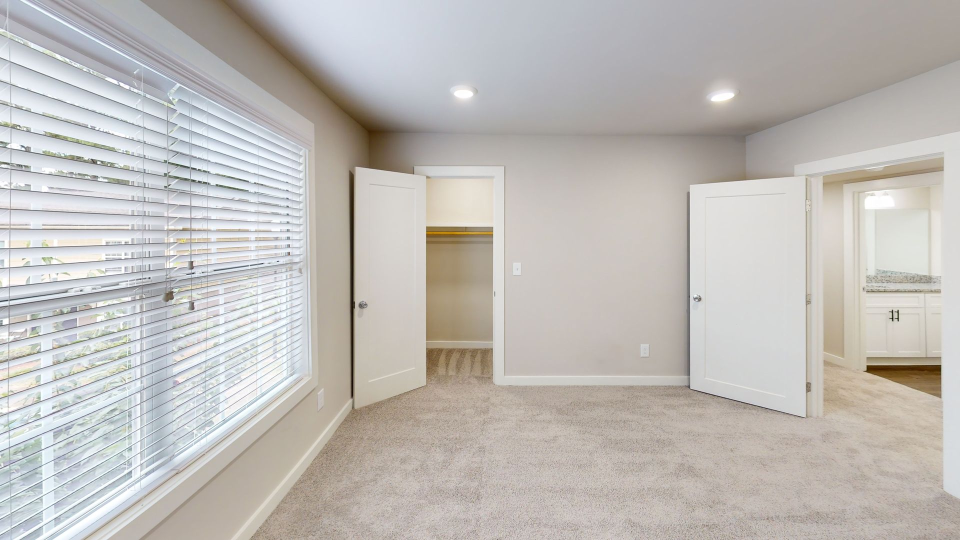 Empty beige bedroom with carpet, large window blinds, and open closet and bathroom doors.