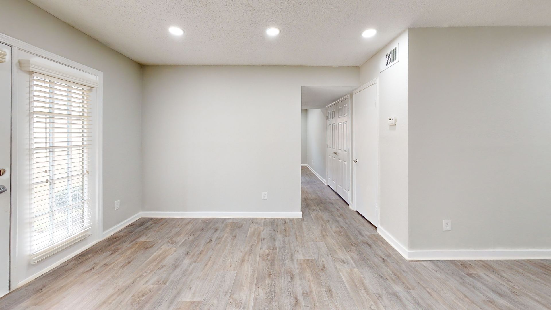 Bright empty living room with light wood floors, recessed lights, and a hallway opening to the right.