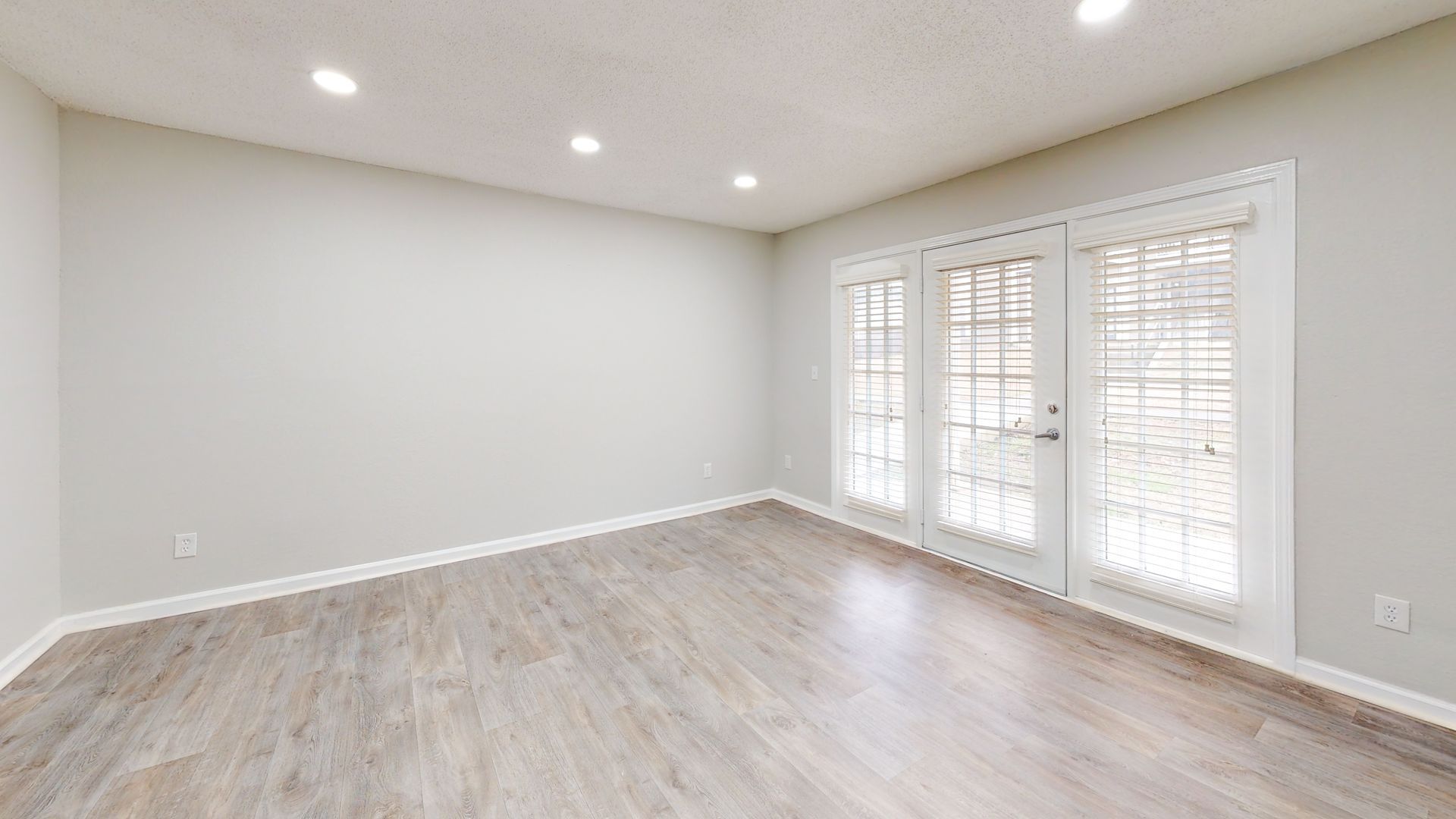 Empty bright living room with light wood floors, white walls, and French doors with blinds