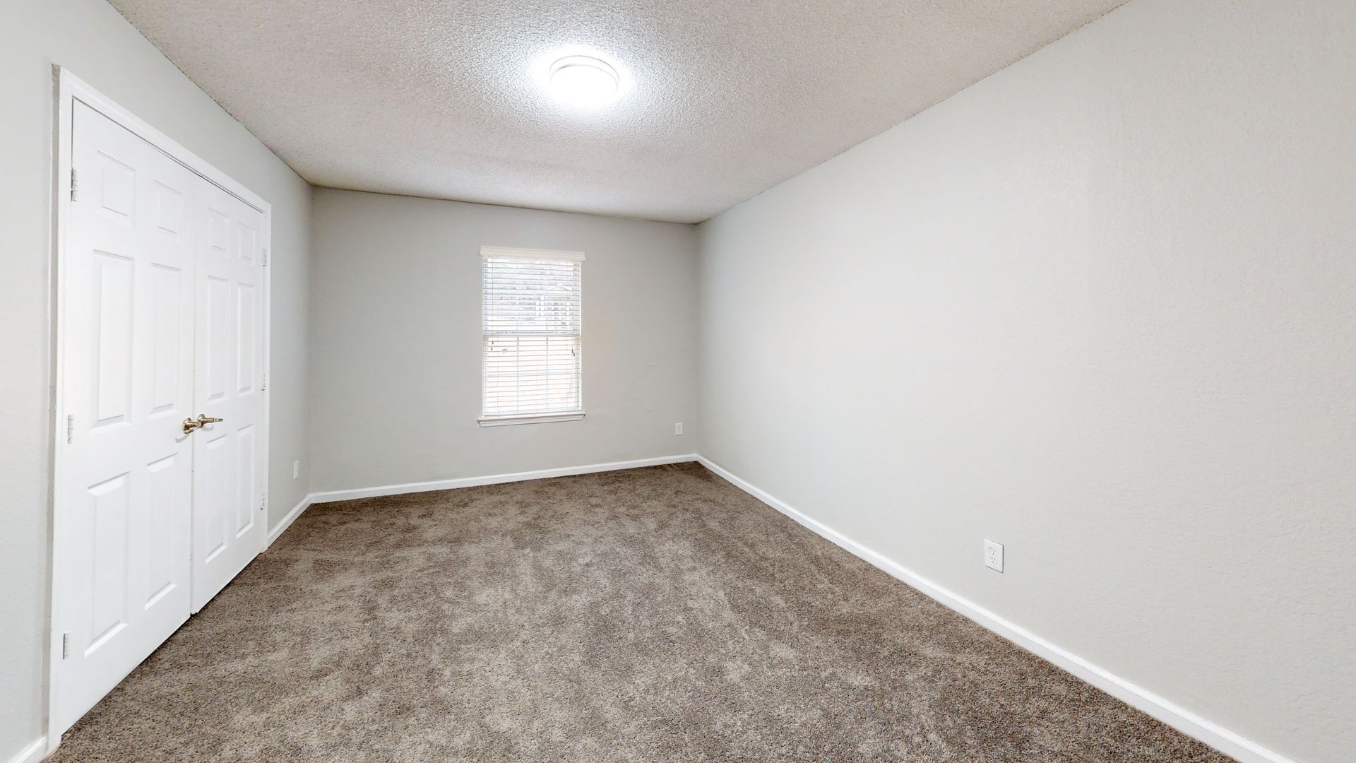 Empty carpeted bedroom with white walls, a window, and double closet doors.