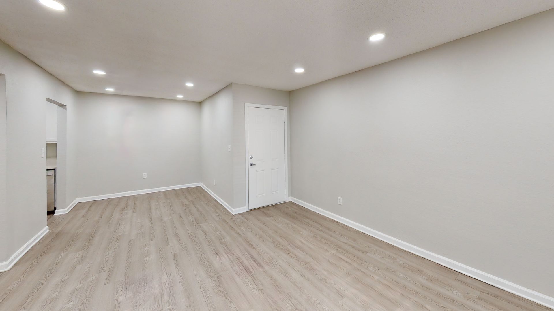 Empty white room with light wood floor, recessed lights, and a closed door