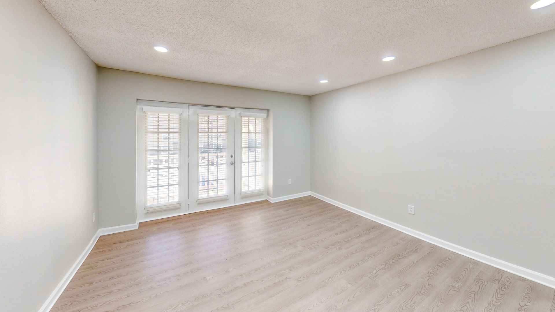 Empty beige living room with light wood floor, recessed lights, and French doors leading outside