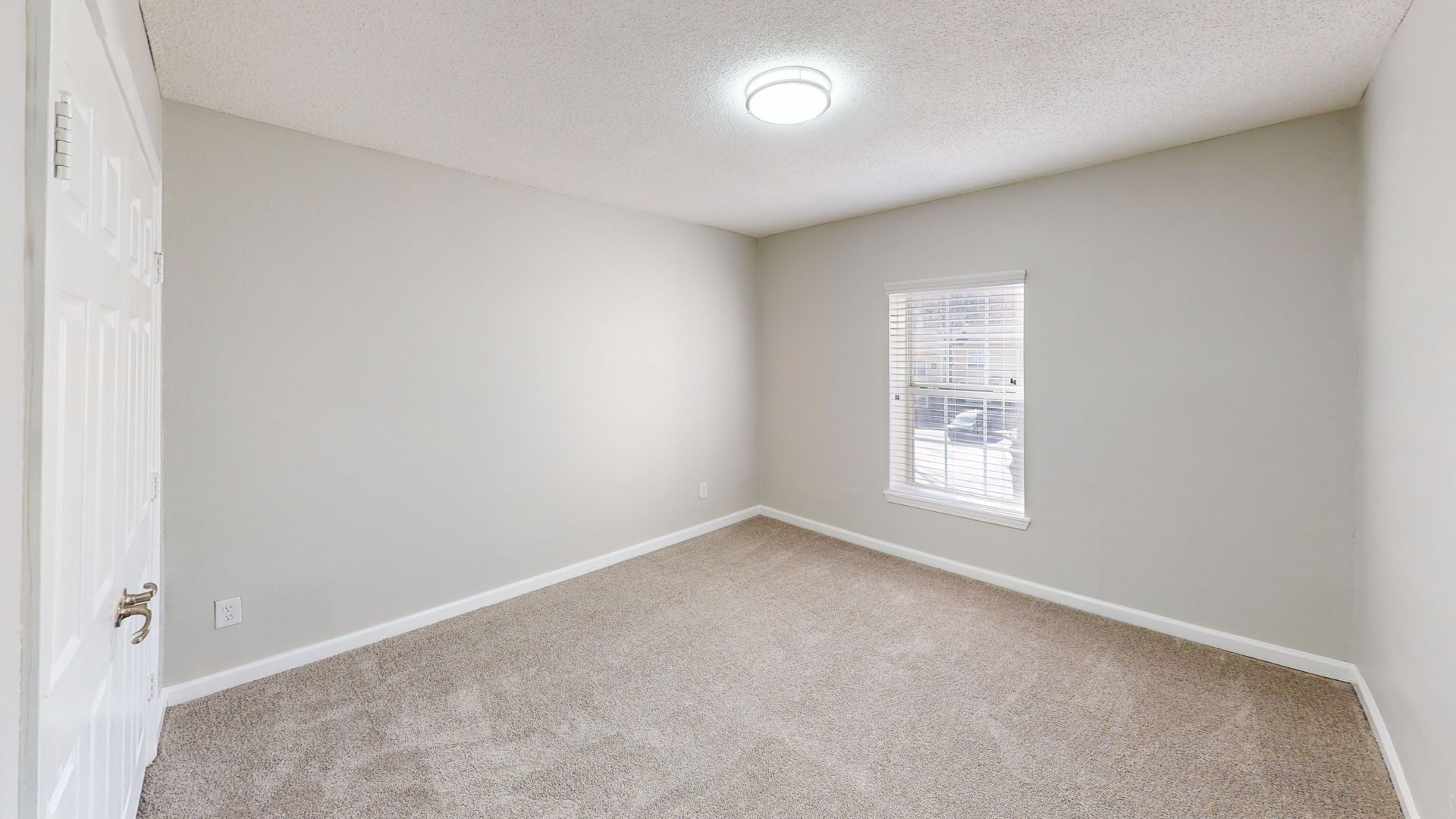 Empty beige carpeted bedroom with white walls, a window, and a ceiling light