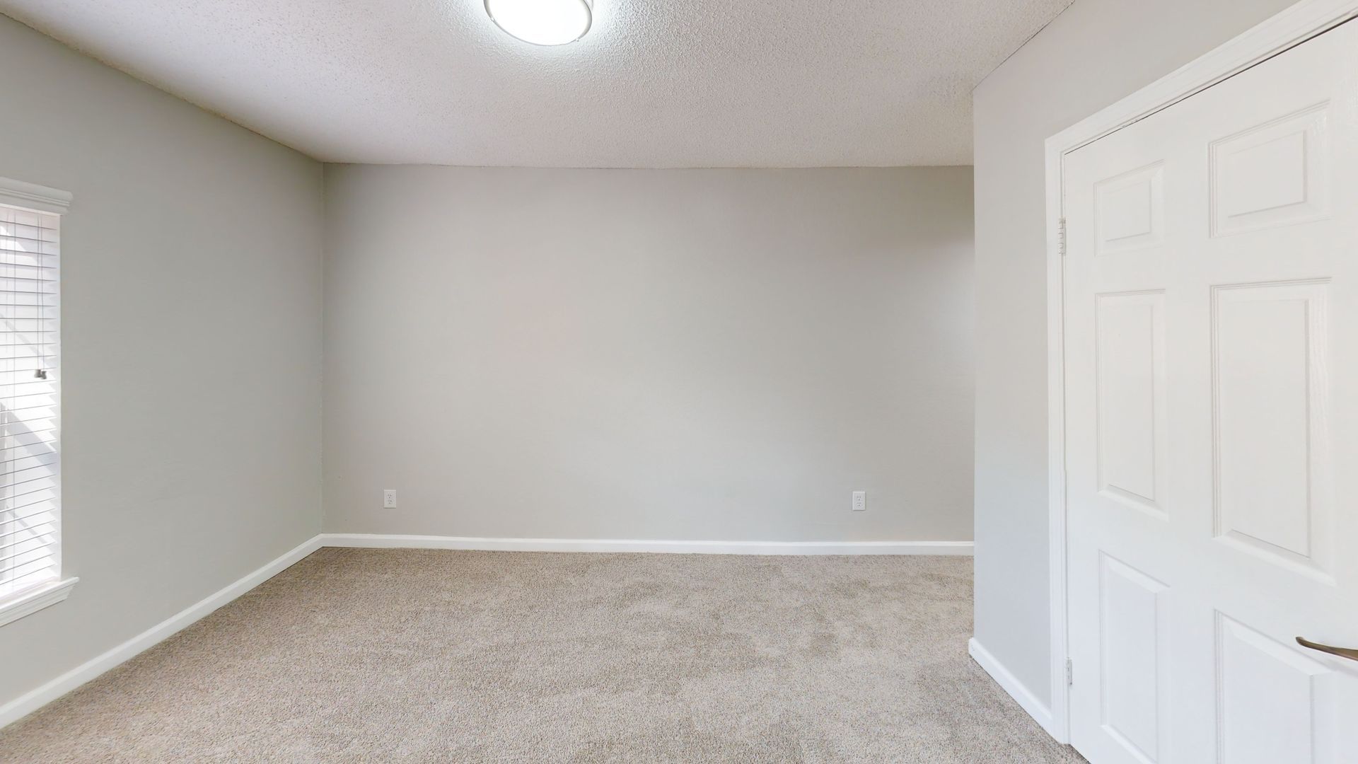 Empty beige bedroom with carpet, white closet doors, and a window on the left.