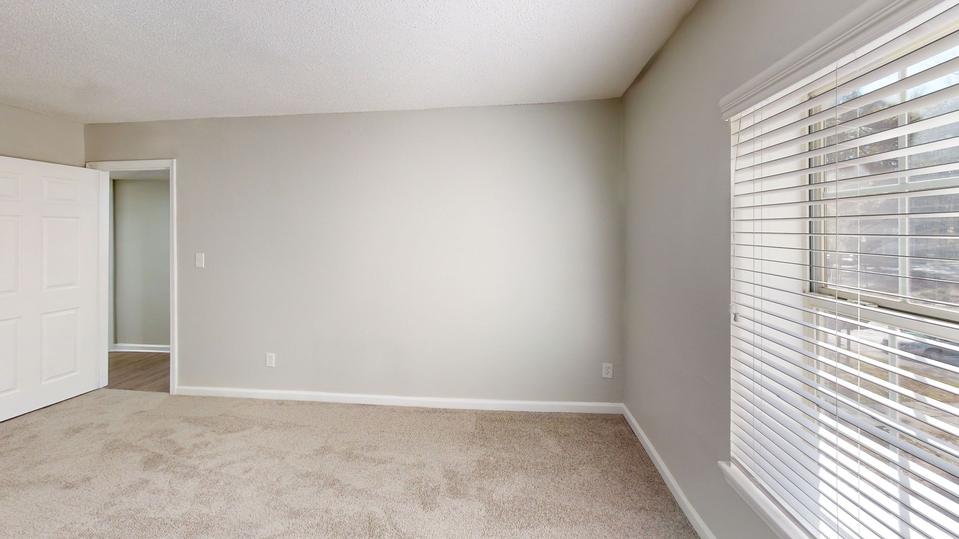 Empty beige bedroom with carpet, white walls, open closet door, and window blinds.