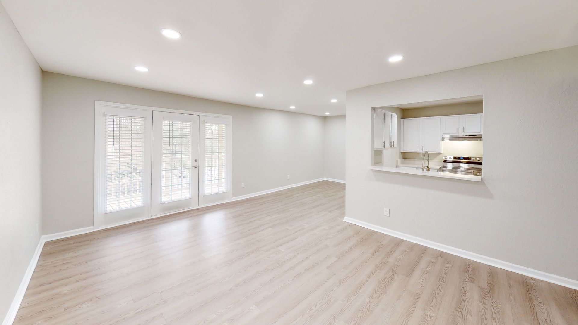 Bright empty living room with light wood floors, recessed lights, and pass-through to kitchen