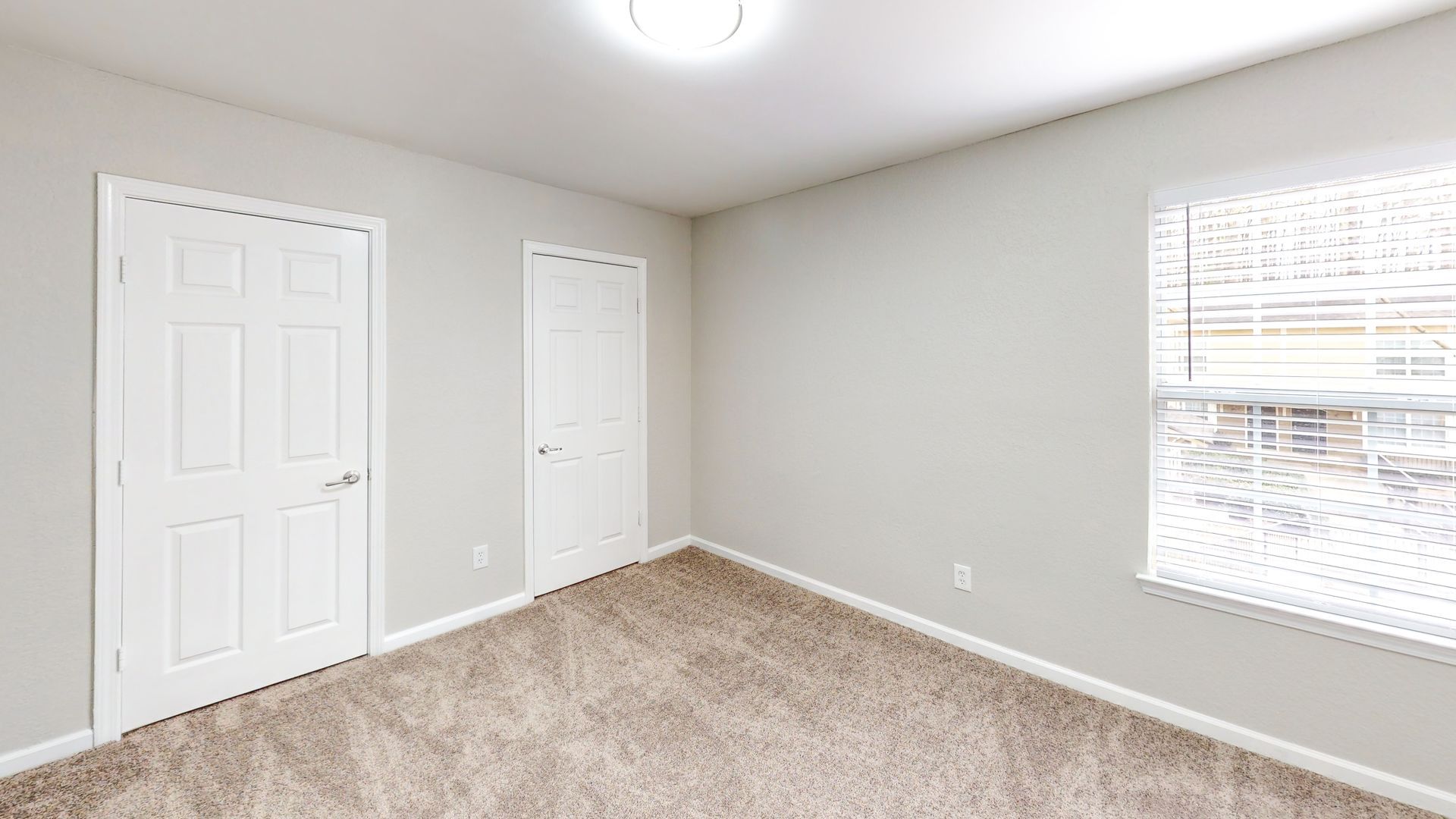 Empty beige bedroom with carpet, two white closet doors, and a window with blinds.