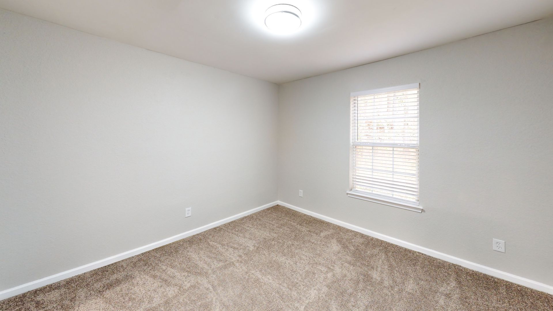 Empty beige bedroom with carpet, white walls, ceiling light, and a small window with blinds