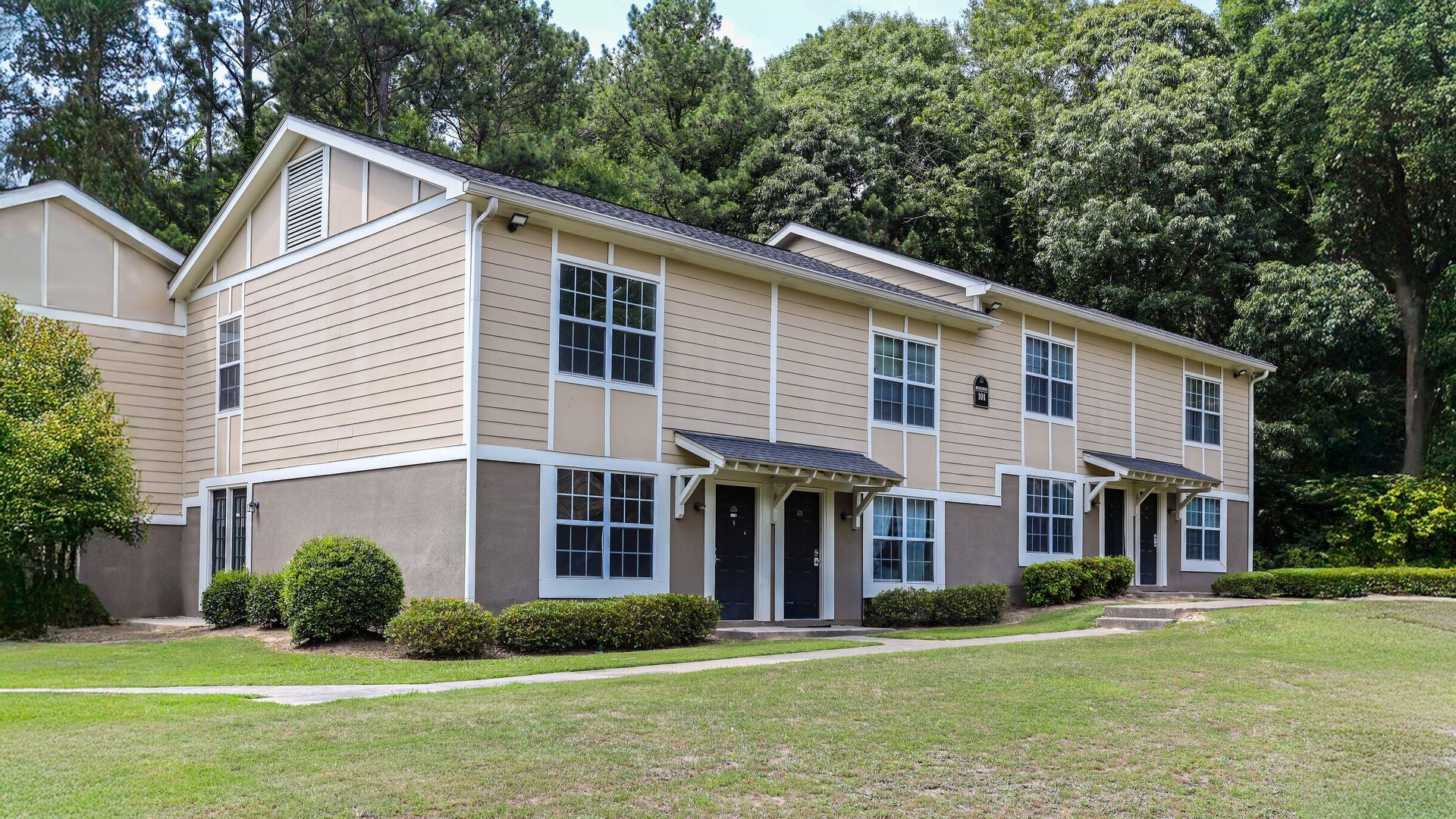 Two-story beige apartment building with white trim, black shutters, and a grassy lawn surrounded by trees