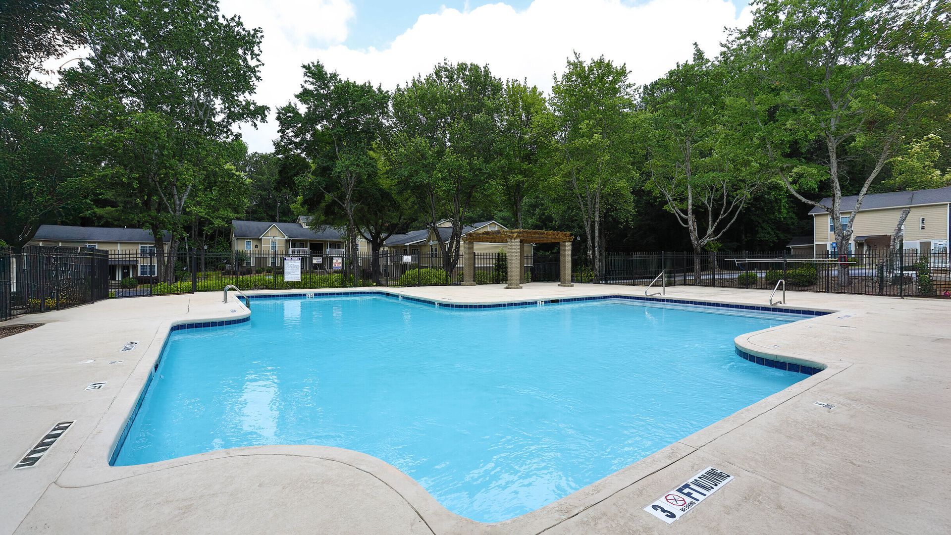 Outdoor swimming pool with blue water, surrounded by a concrete deck and trees.