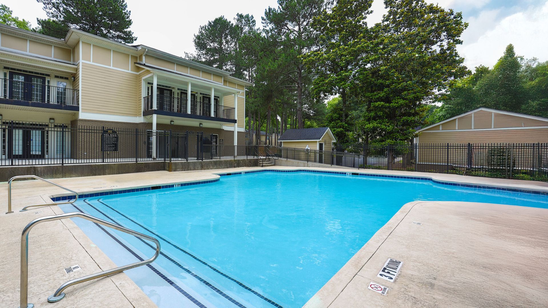 Outdoor swimming pool beside a beige clubhouse and trees, with a fenced deck and clear blue water.