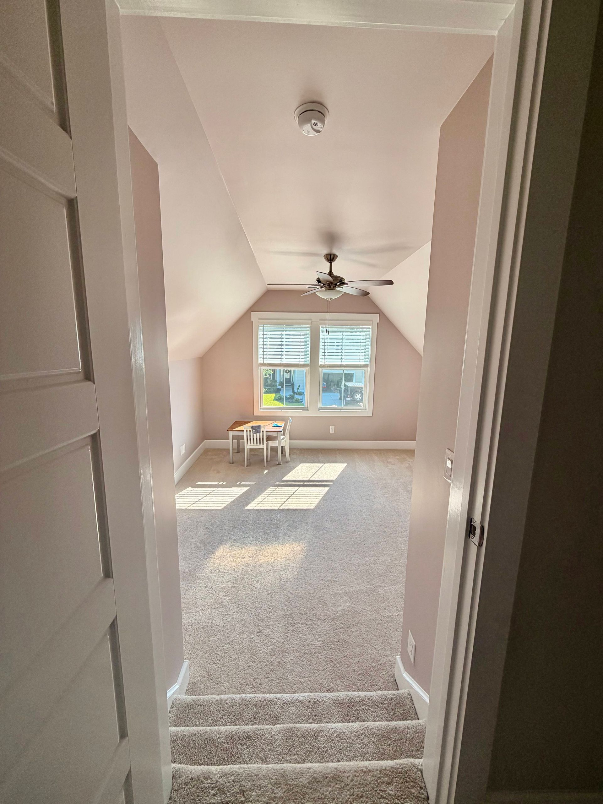 Empty carpeted attic room with sloped ceiling, ceiling fan, and sunlight from a small window.