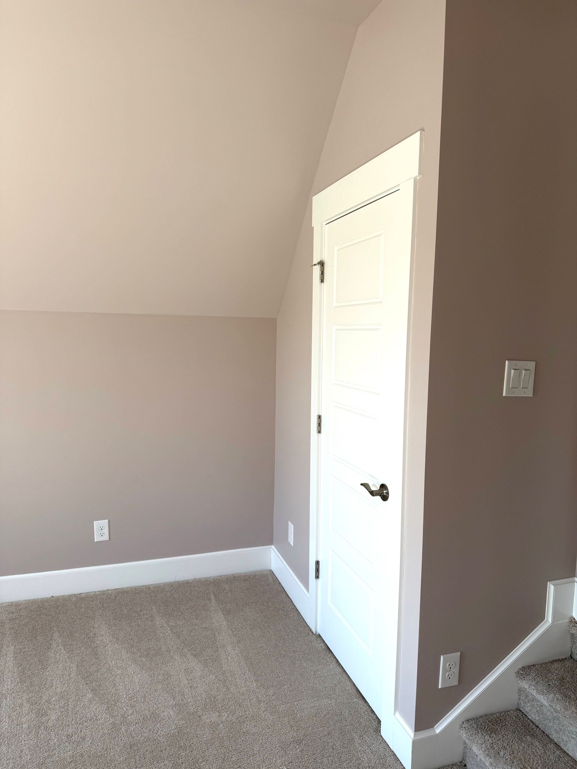 Small empty carpeted room with taupe walls, white trim, and a closed white door by stairs